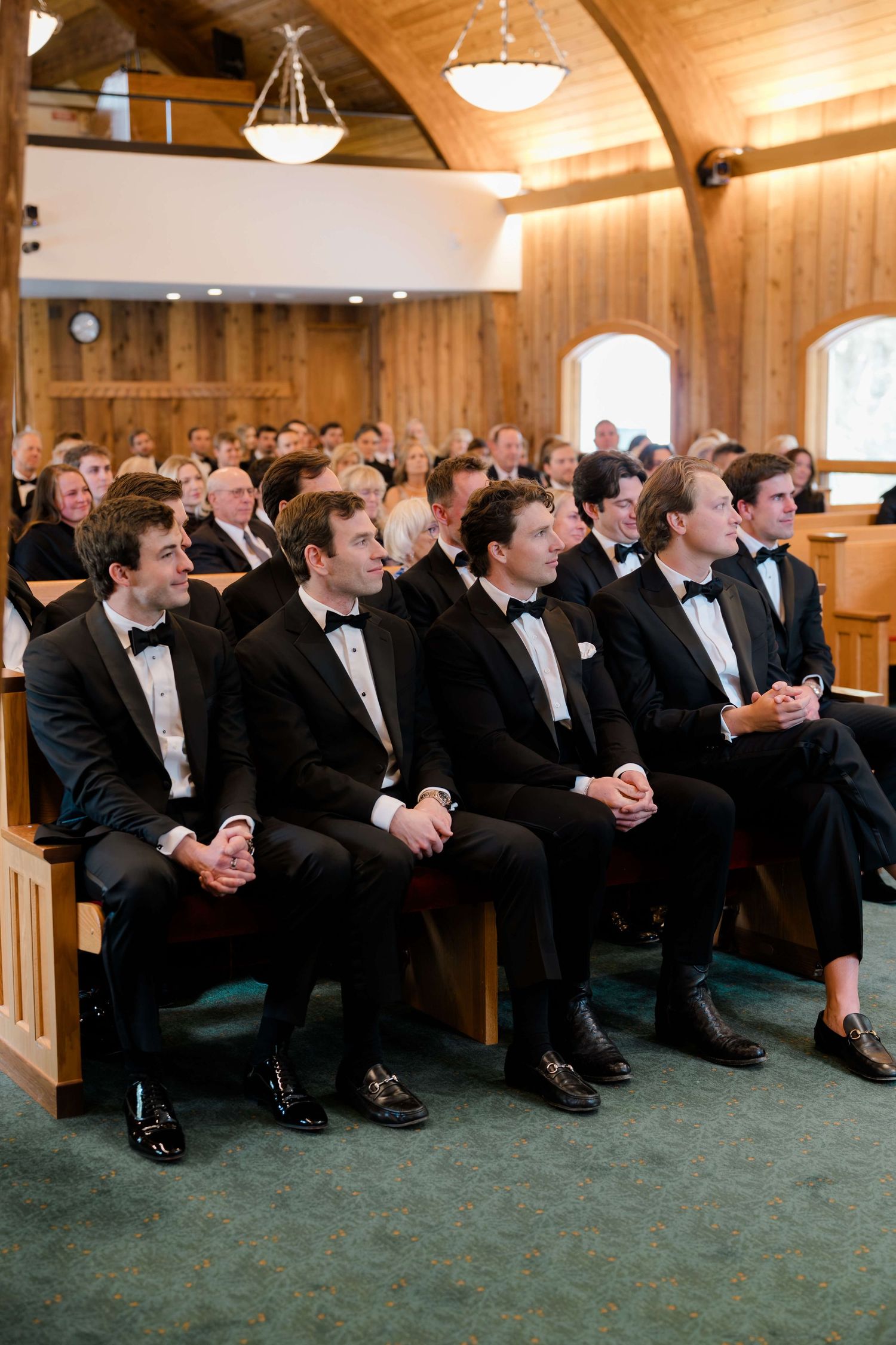 Groomsmen in black tuxedos seated in wooden church pews during a wedding ceremony.