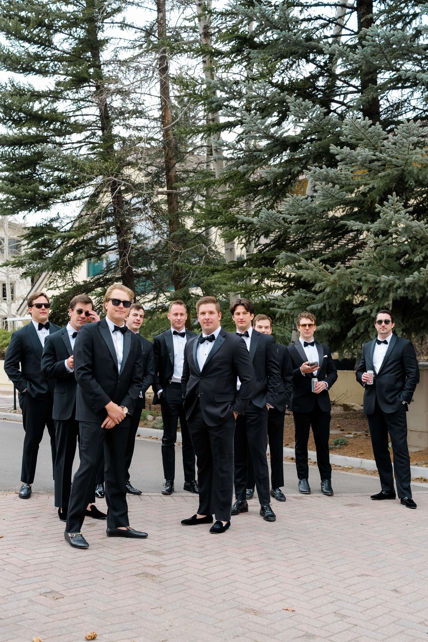 Groomsmen in black tuxedos and bow ties posing outdoors with evergreen trees backdrop at a wedding.