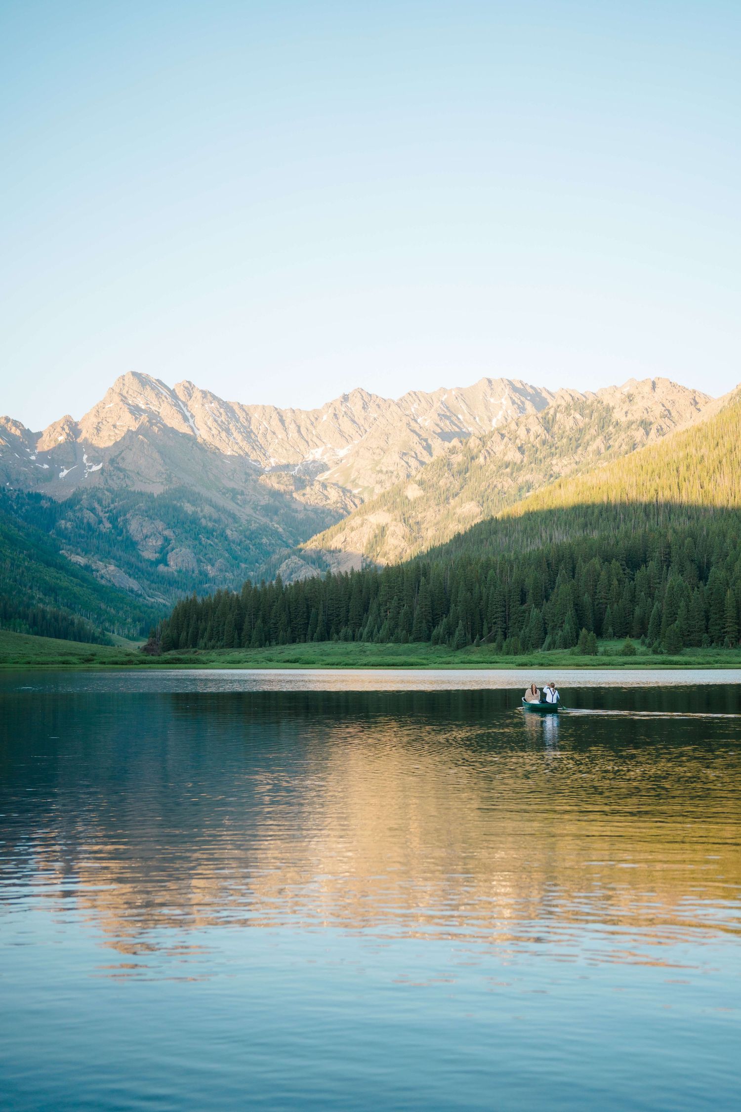 A mountain lake reflects peaks and pine forests at sunset, with a small boat gliding across the calm water at Piney River Ranch Wedding.
