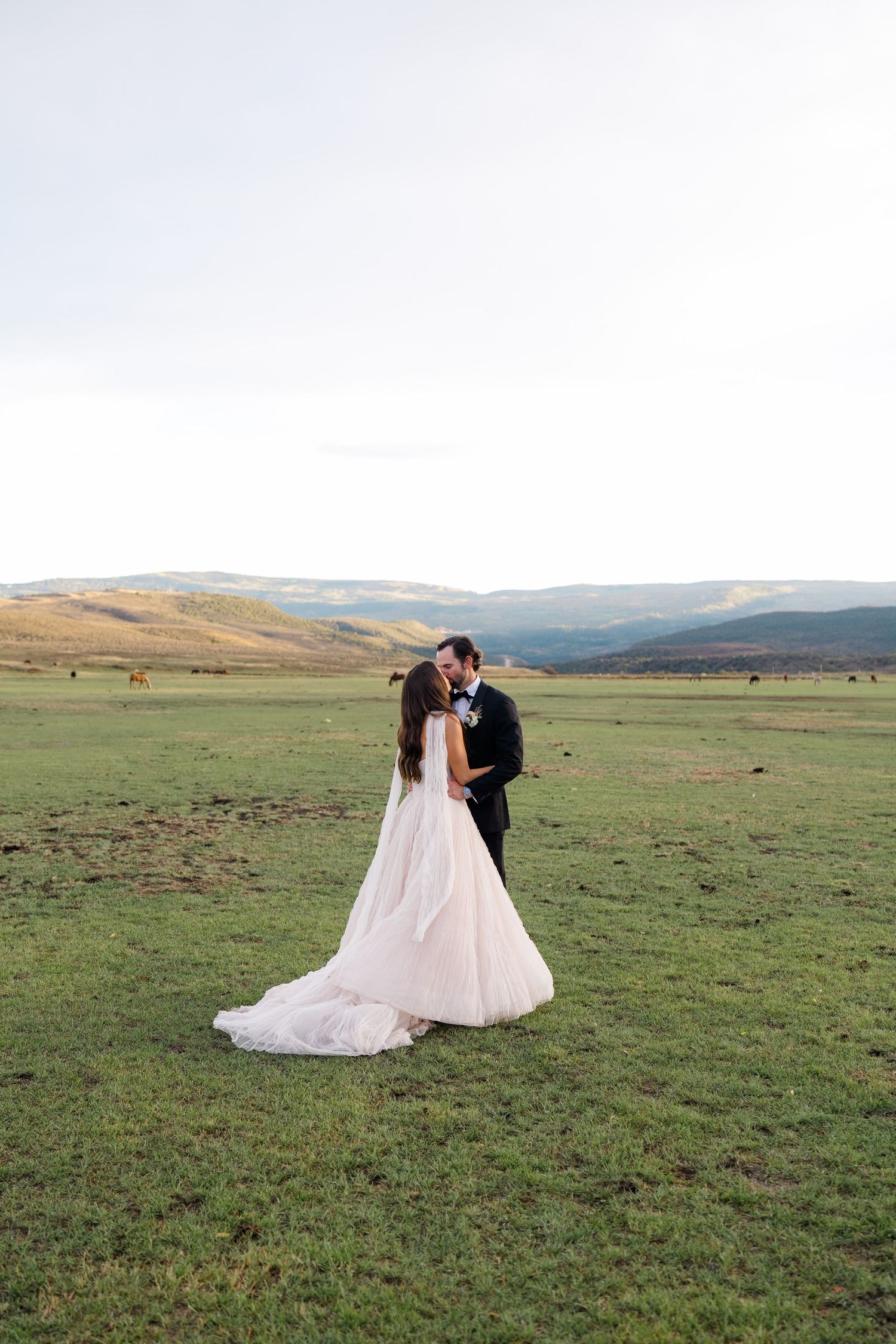 Bride and groom share a kiss in a vast green meadow with rolling mountains in the background on their wedding day.