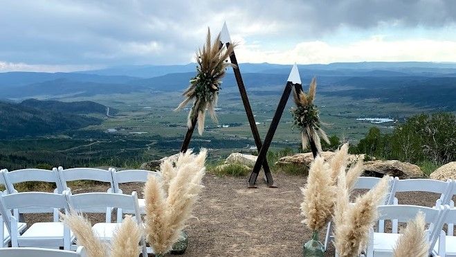 Outdoor mountain wedding ceremony setup with triangular arch, pampas grass, white chairs, and panoramic valley views.