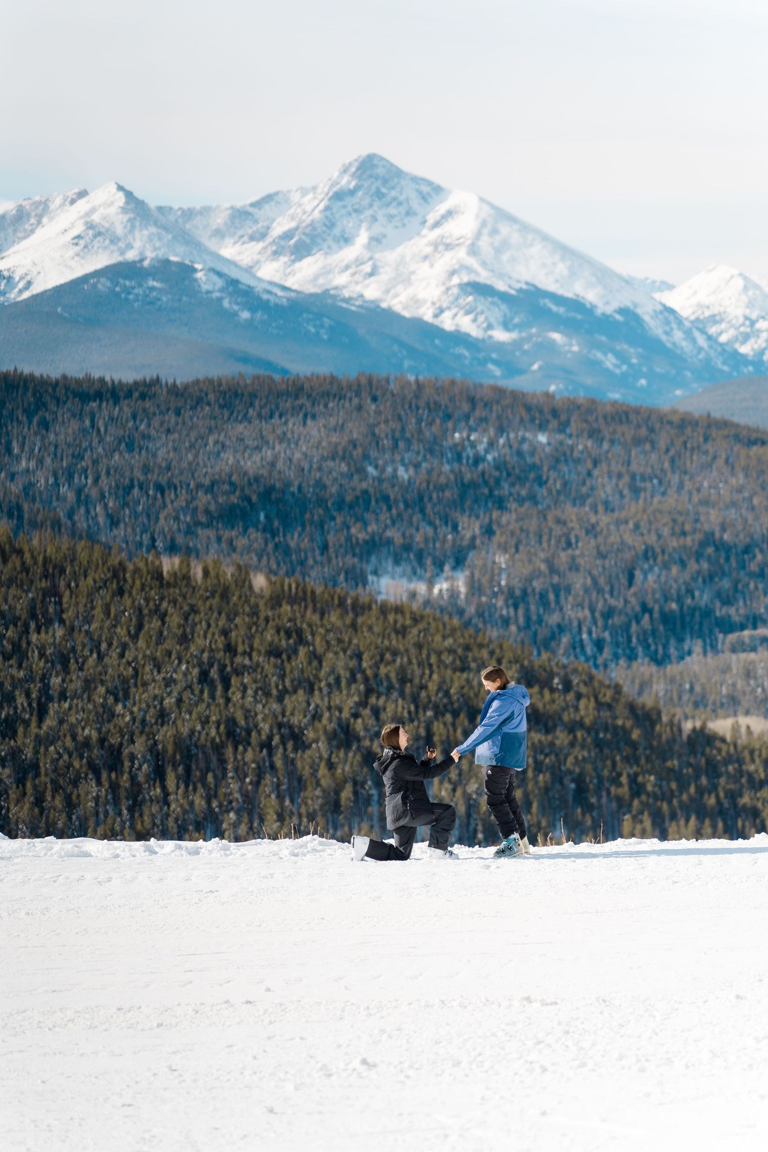 Two people on a snowy mountain slope with majestic snow-capped peaks and evergreen forest in the background.