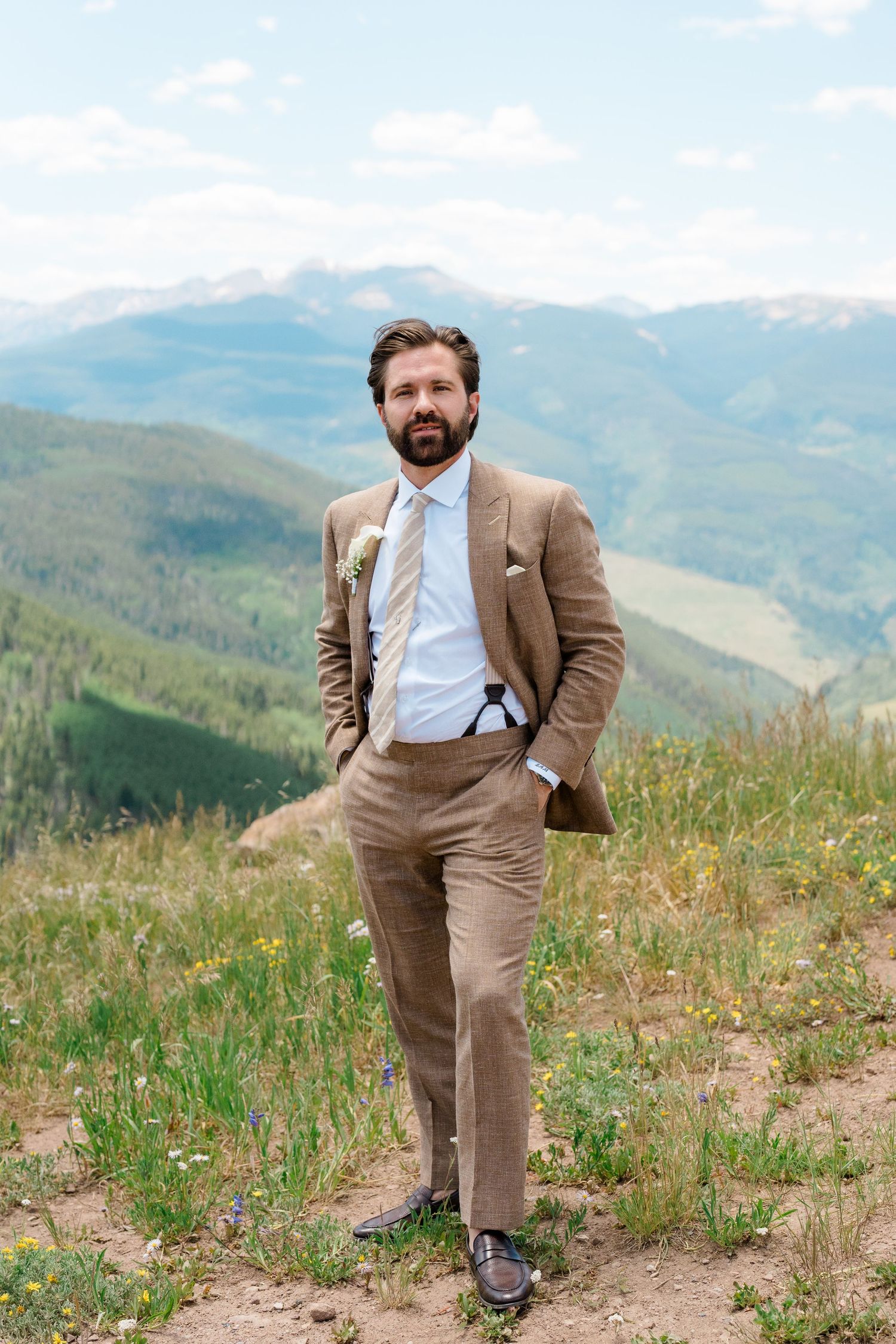 Groom in tan suit stands confidently on mountain overlook with scenic valley views.