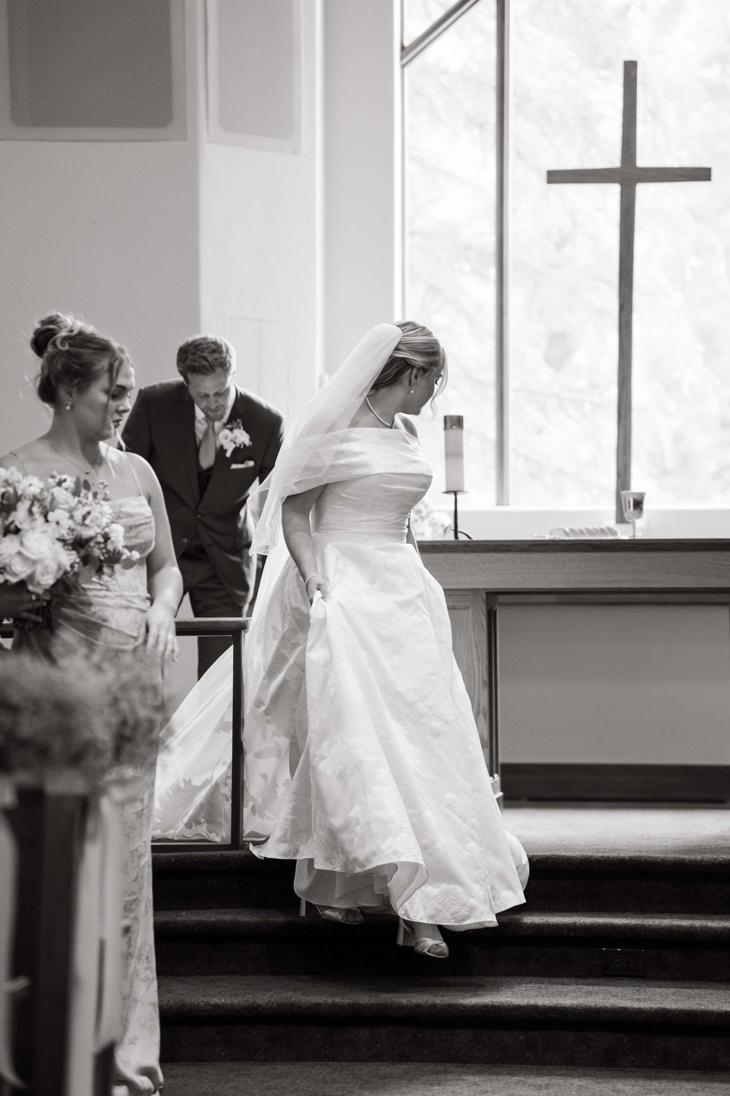Black and white photo of a bride ascending church steps in a flowing white gown with a cross visible in the background at vail wedding.