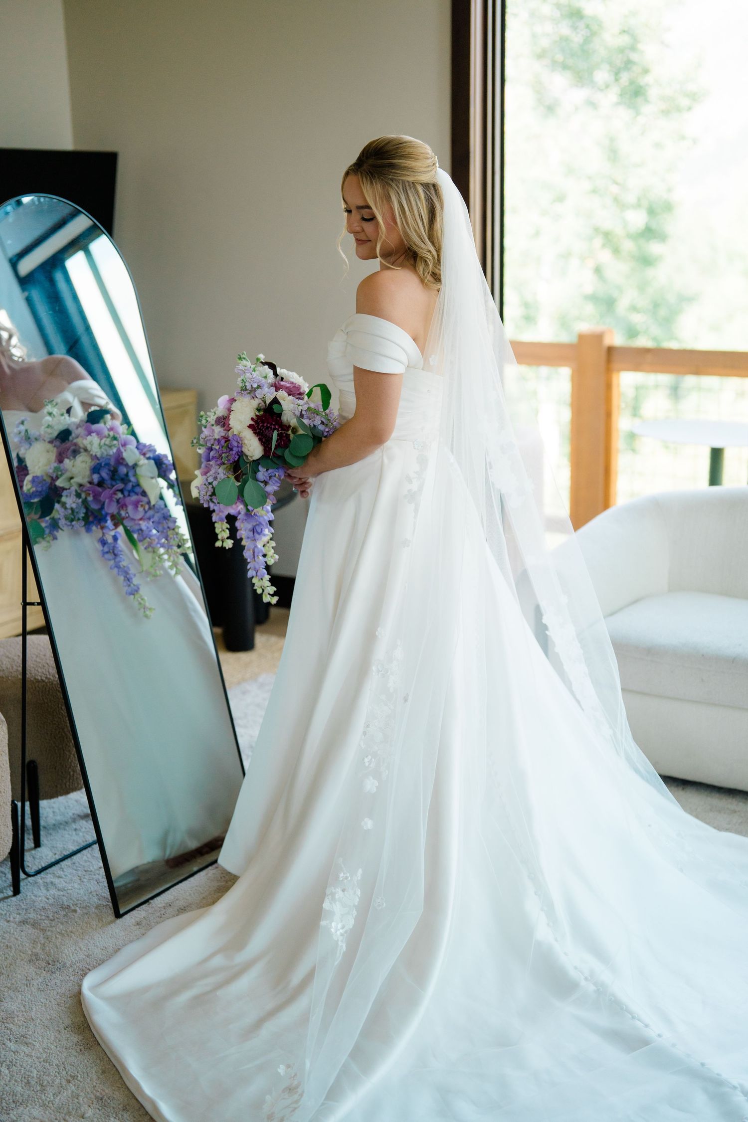 A bride in a flowing white wedding gown stands before a mirror holding a purple and blue floral bouquet during Breckenridge wedding.
