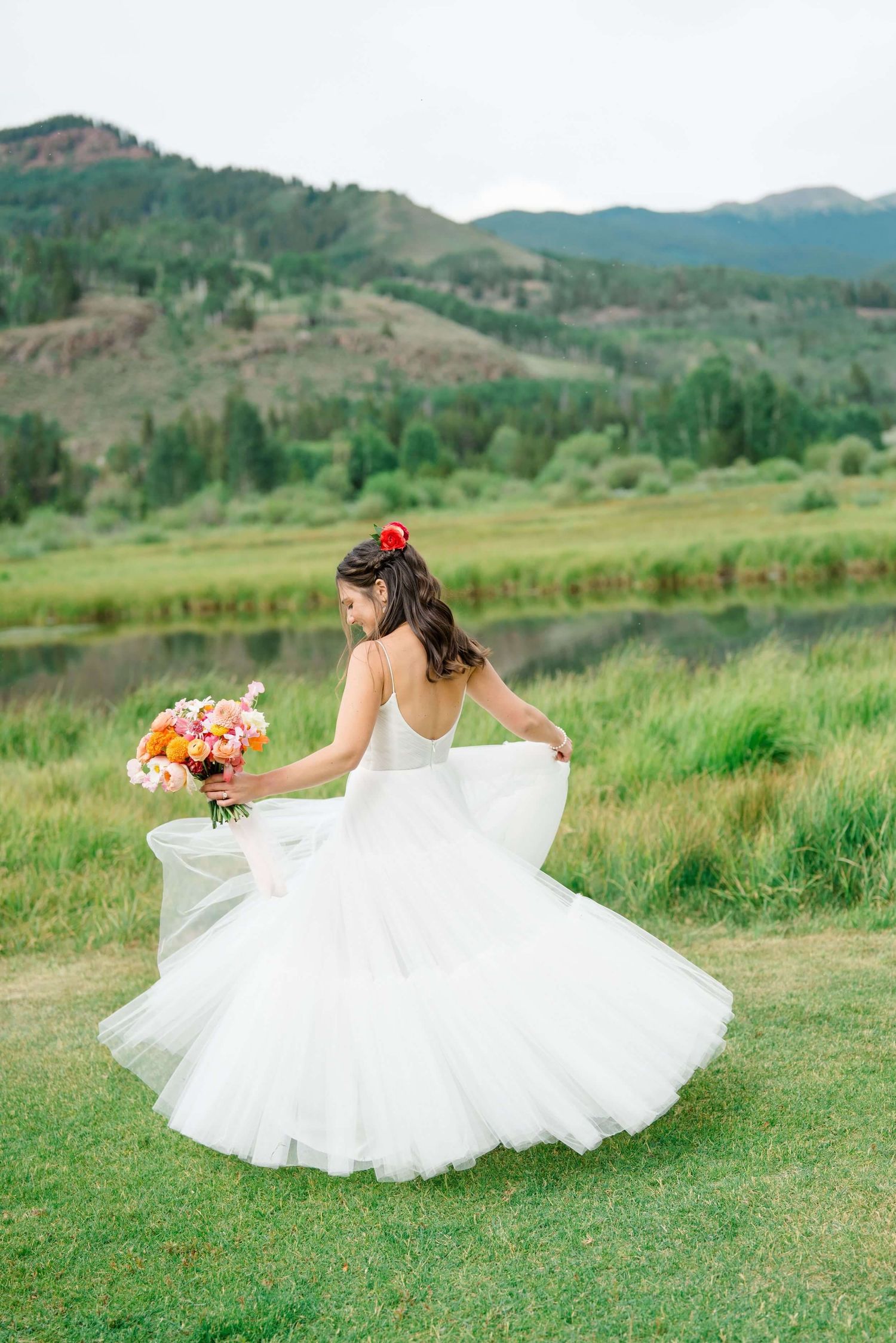 Bride in flowing white dress twirling on a grassy hillside with mountain landscape in background at Camp Hale.