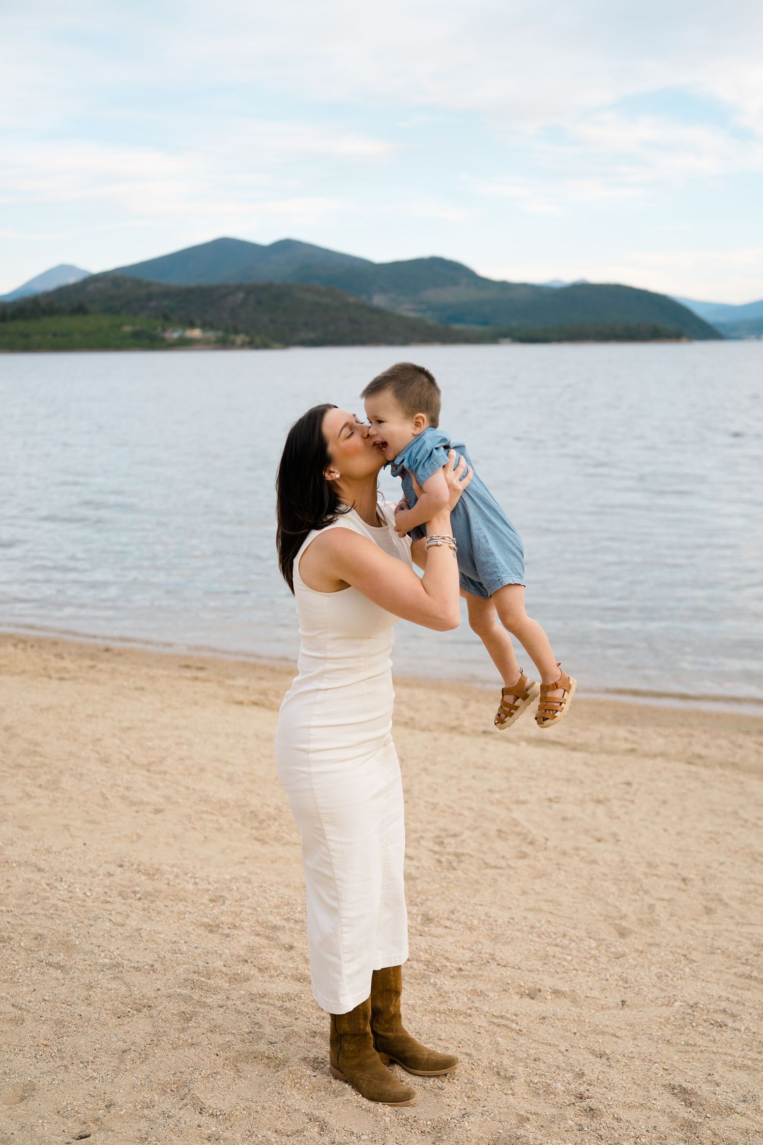 A parent in a white dress lifts and spins a small child wearing blue on a beach with mountains in the background at lake dillon.