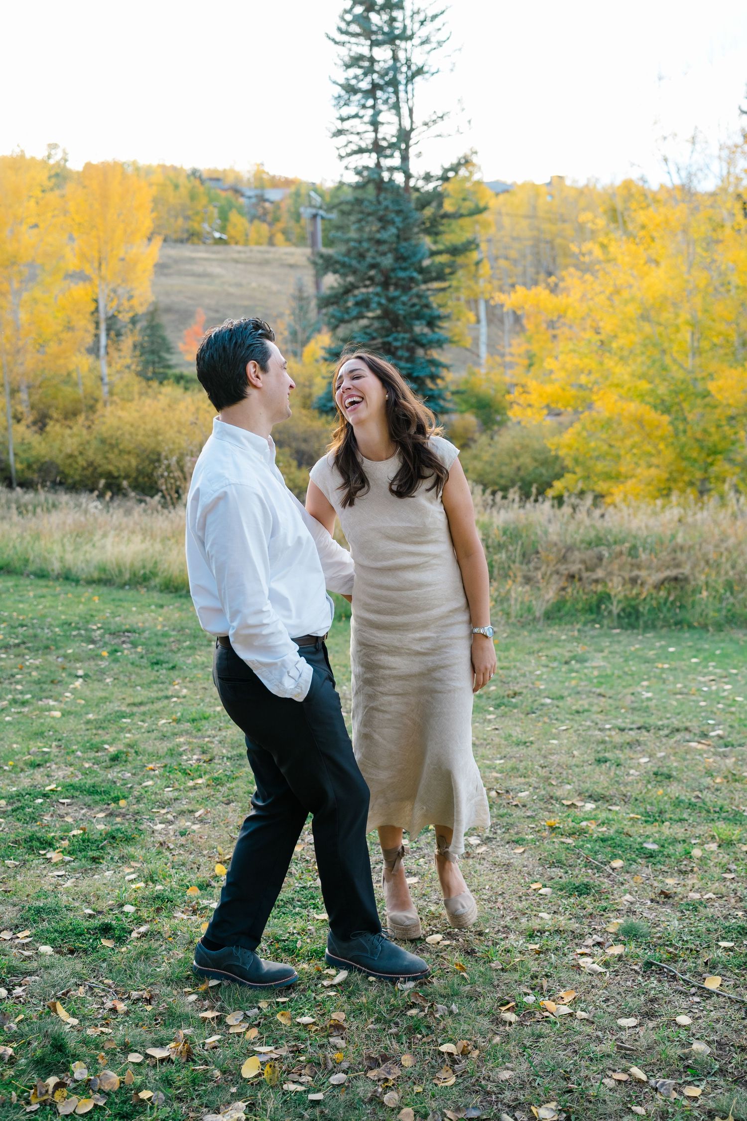 A couple shares playful moments together in a grassy field surrounded by autumn foliage and evergreen trees.