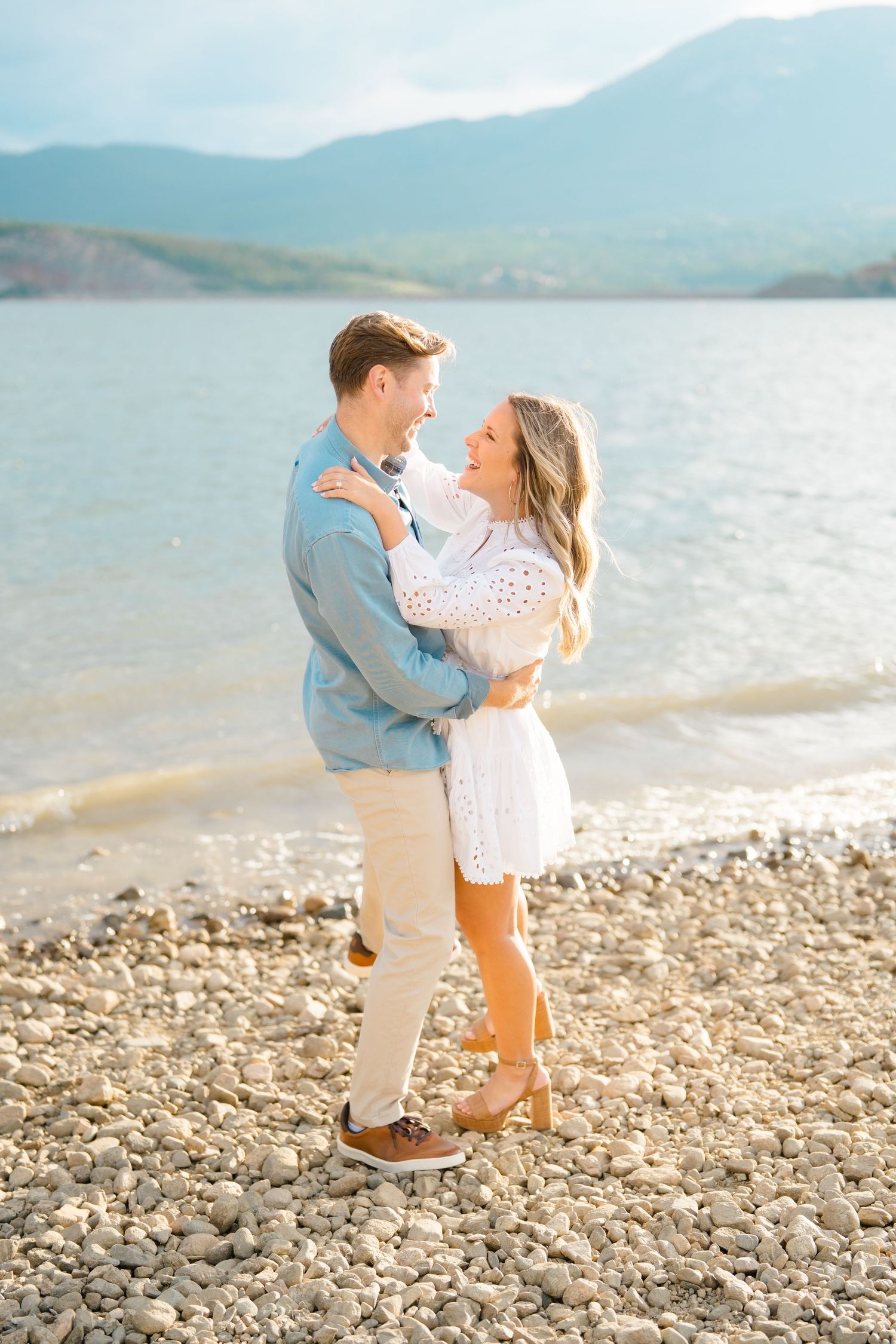 A couple shares a romantic moment on a rocky beach with mountains and water in the background.