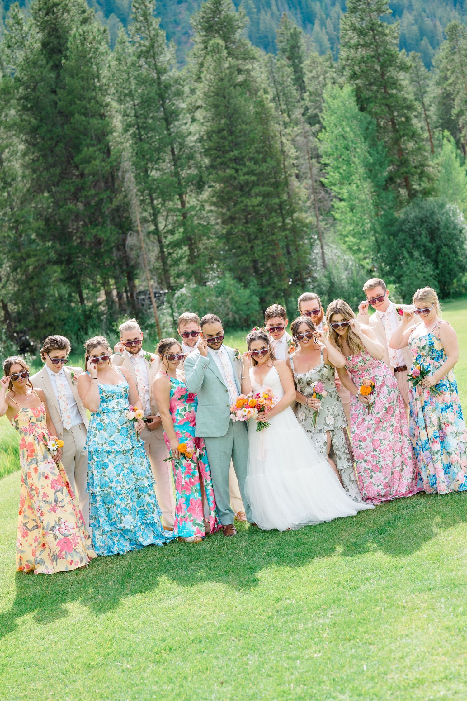 Large wedding party in floral dresses and gray suits posed together in a mountain forest clearing.
