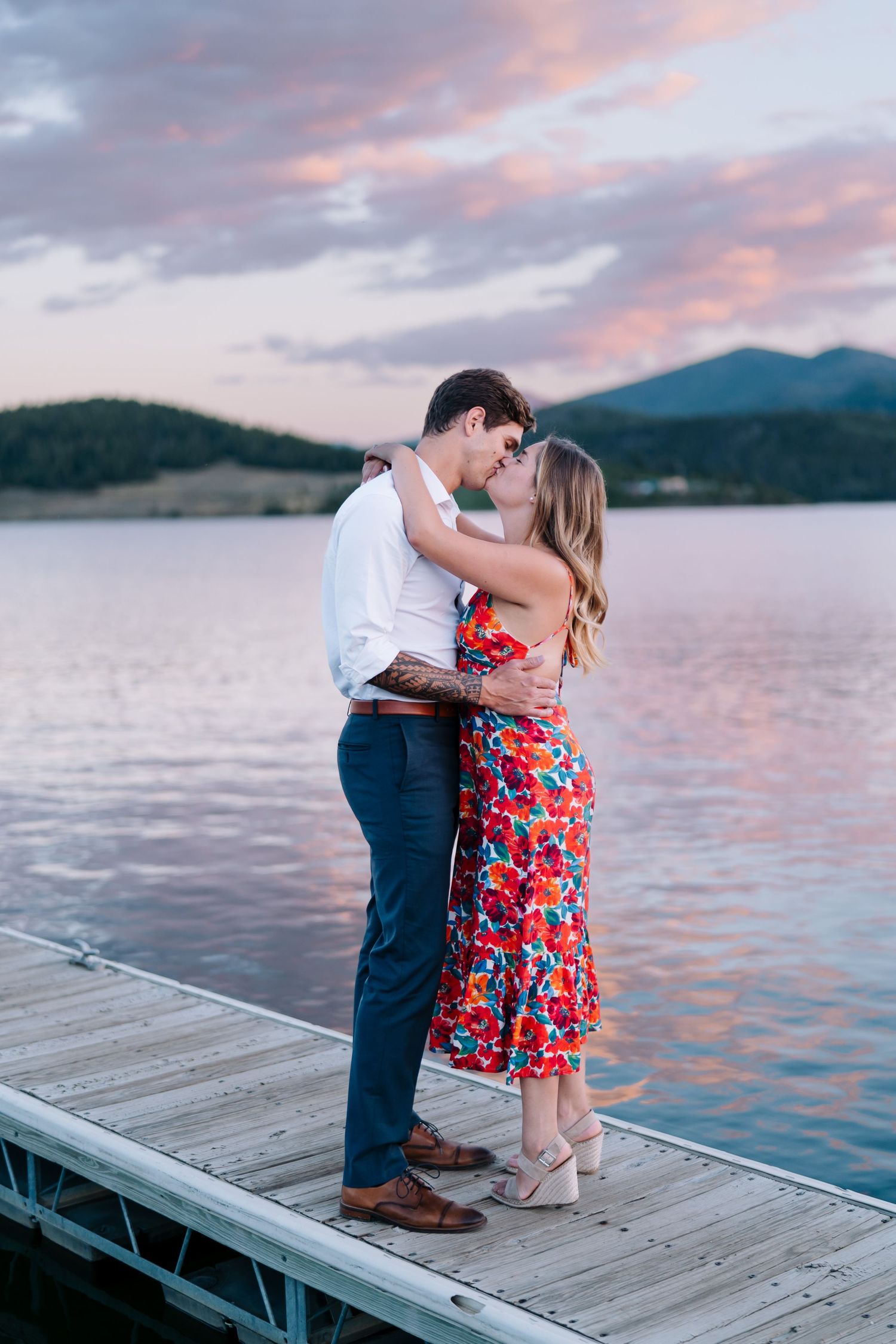 A couple embraces on a wooden dock at sunset with mountains and a lake in the background.