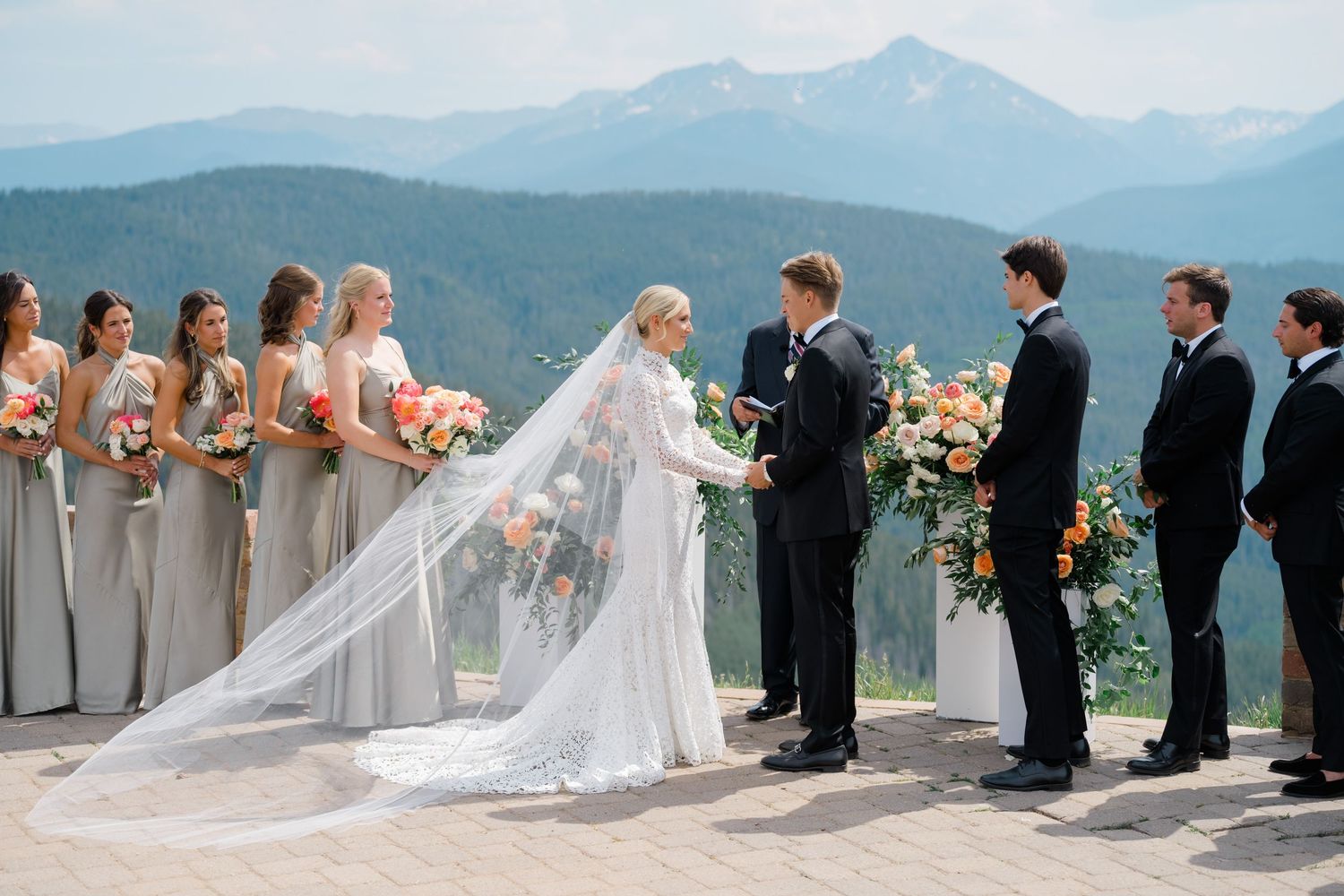 Wedding party stands in line against stunning mountain backdrop with long veil flowing in the breeze at vail mountain wedding deck venue