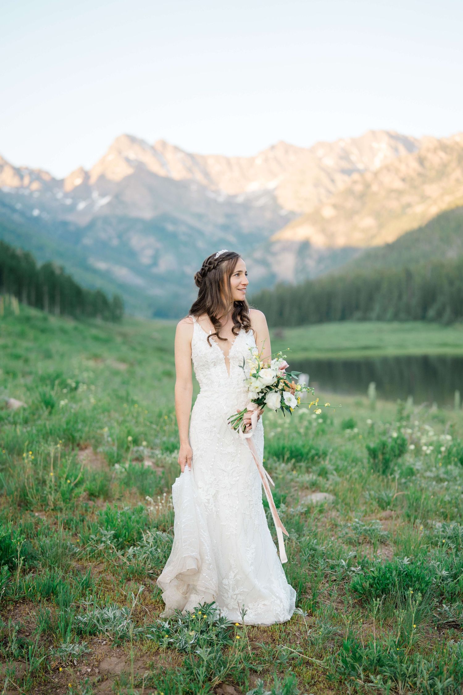 Bride in lace gown holding bouquet stands in mountain meadow with snow-capped peaks and lake at golden hour at Piney River Ranch.