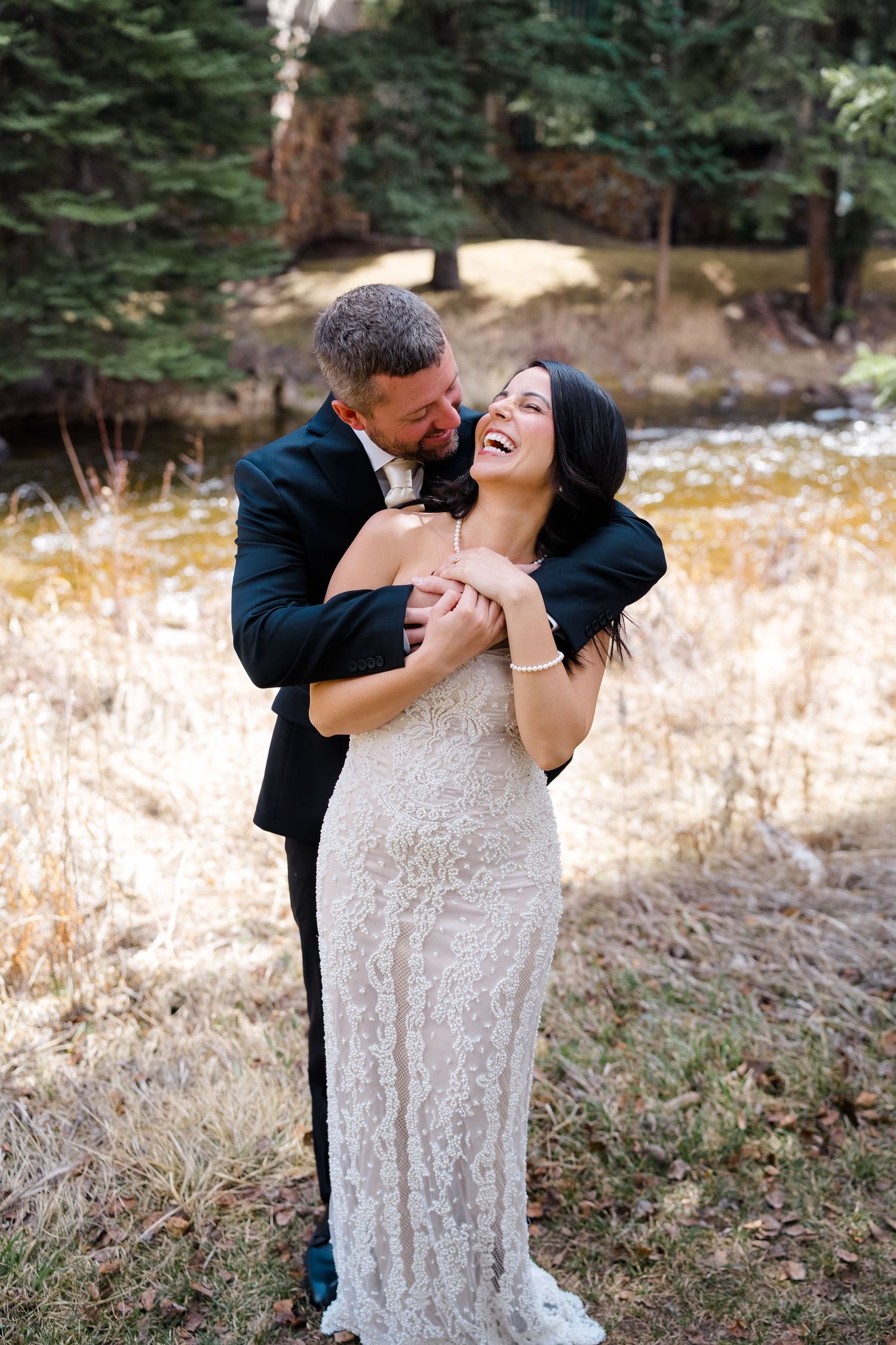 Bride in lace gown laughs as groom in black tuxedo embraces her from behind at their Vail Wedding by Vail Wedding Photographer