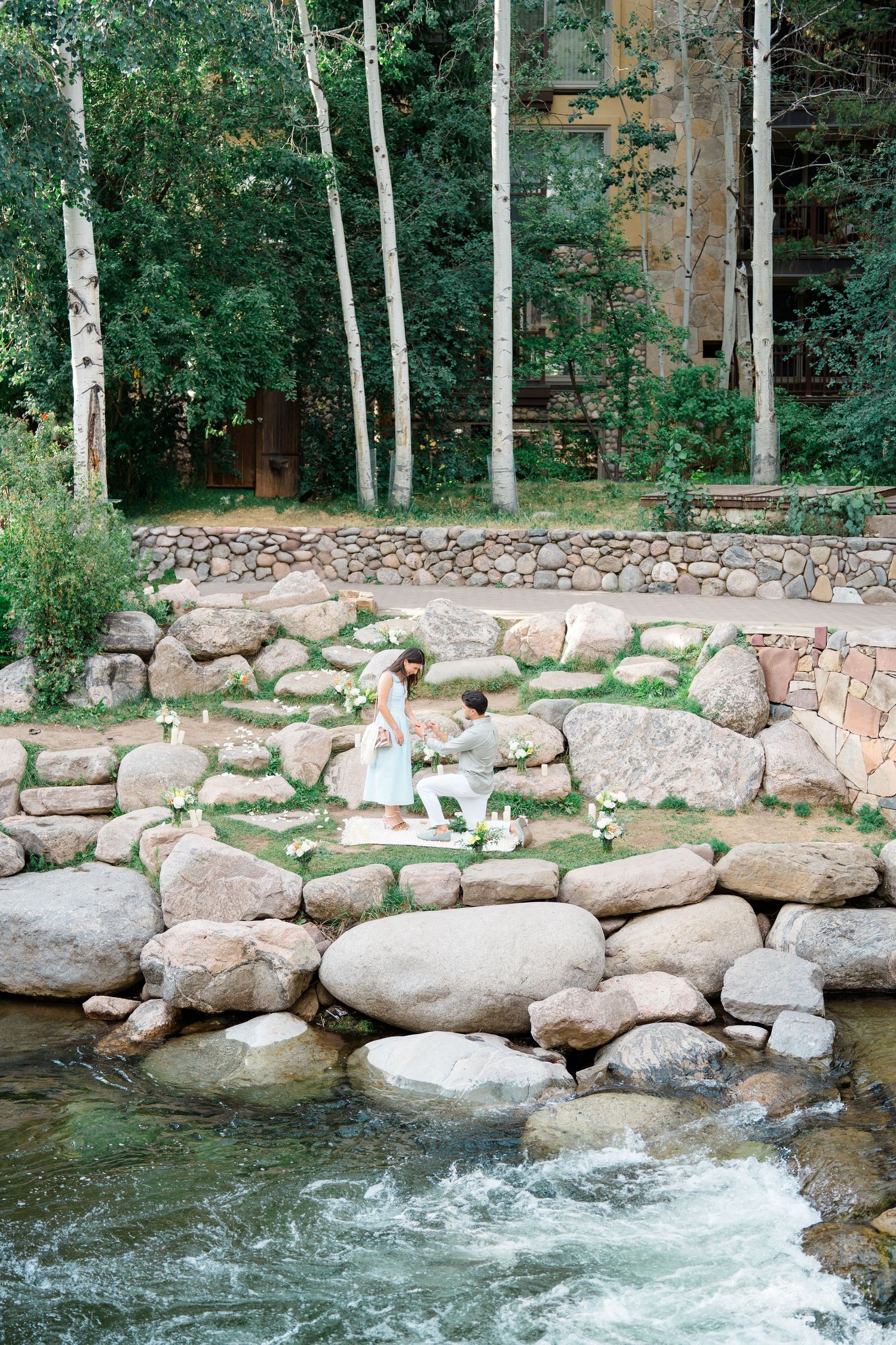A scenic mountain stream flows past stone landscaping and aspen trees in a peaceful outdoor setting.