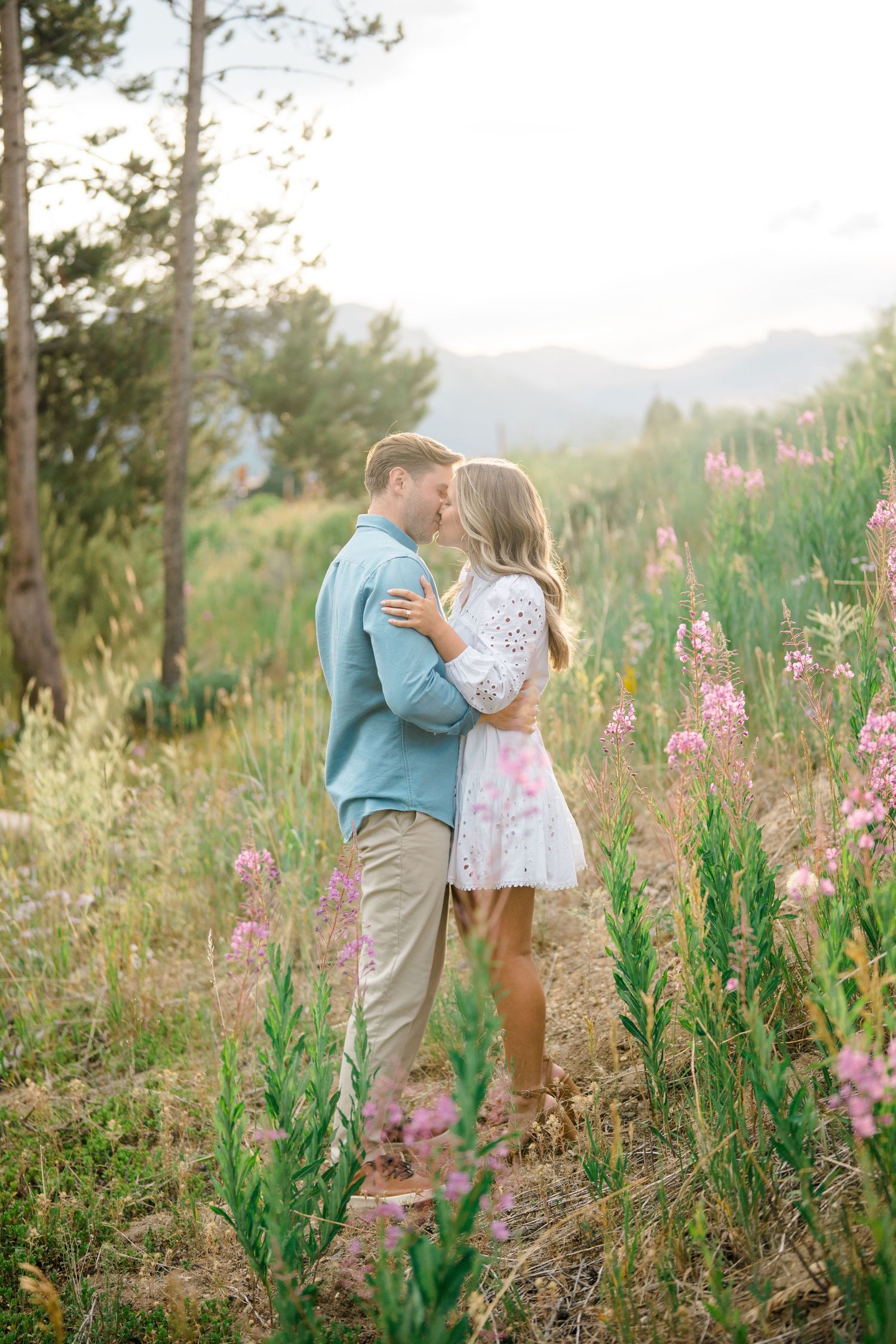 Romantic couple embracing in a field of pink wildflowers with mountains in the distance.
