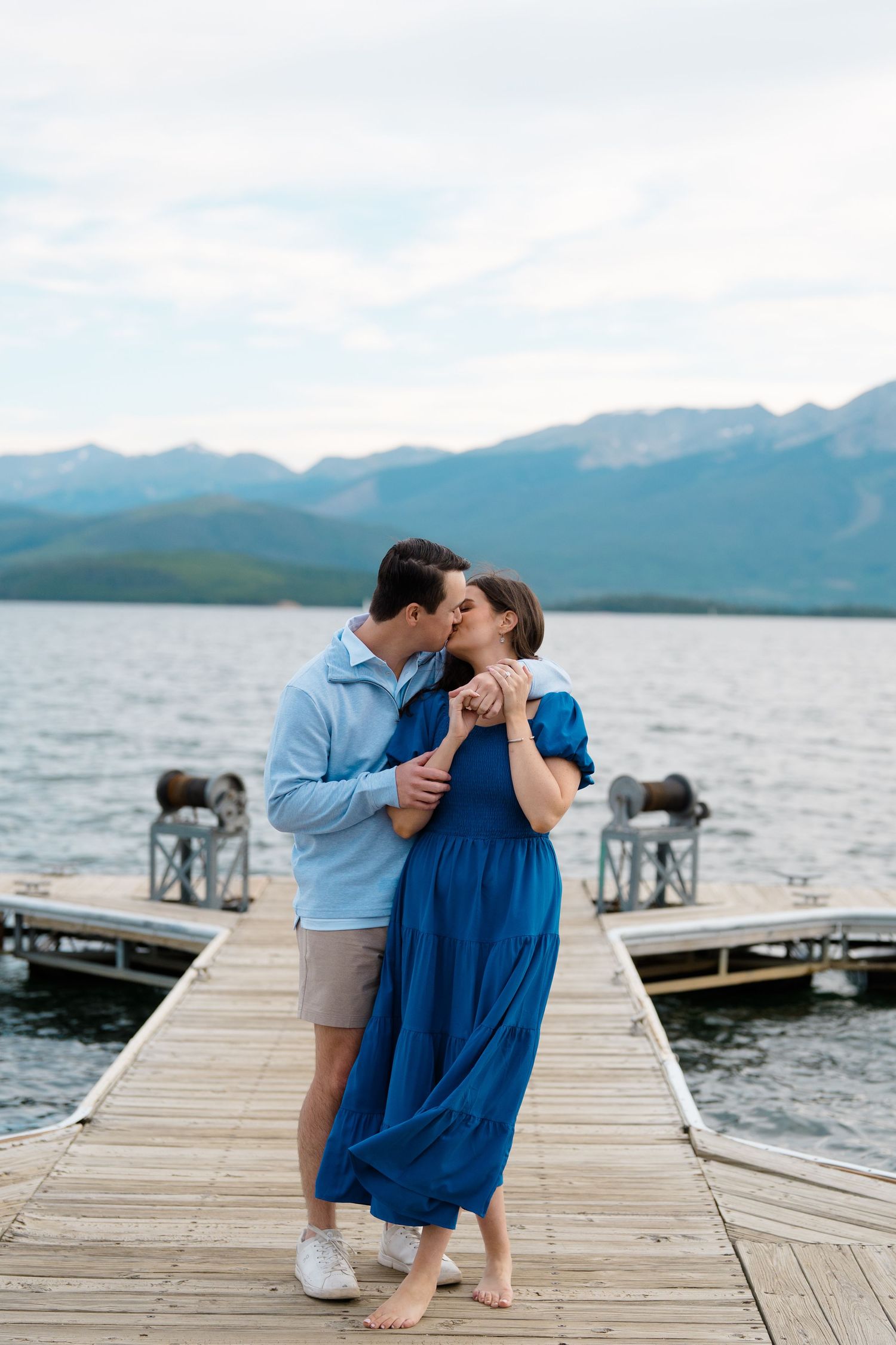 A romantic couple shares intimate moments on a wooden dock overlooking a scenic mountain lake.