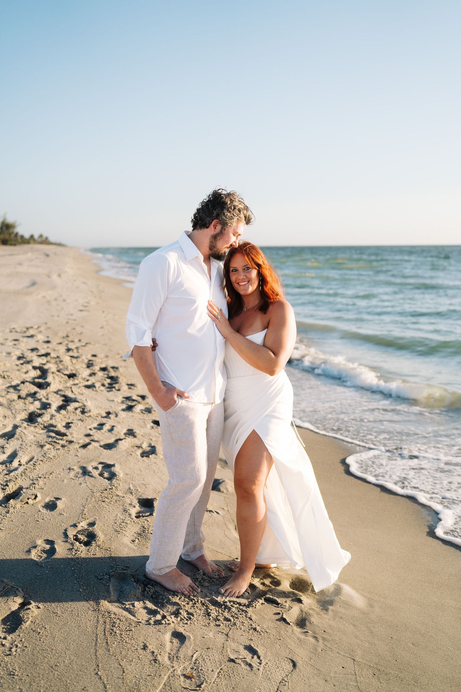 A romantic couple in white attire shares intimate moments during their beach photoshoot at sunset.