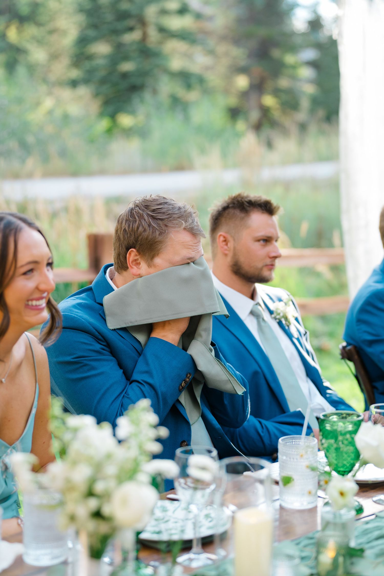 Wedding guests in blue attire seated at an outdoor reception table with white floral centerpieces.
