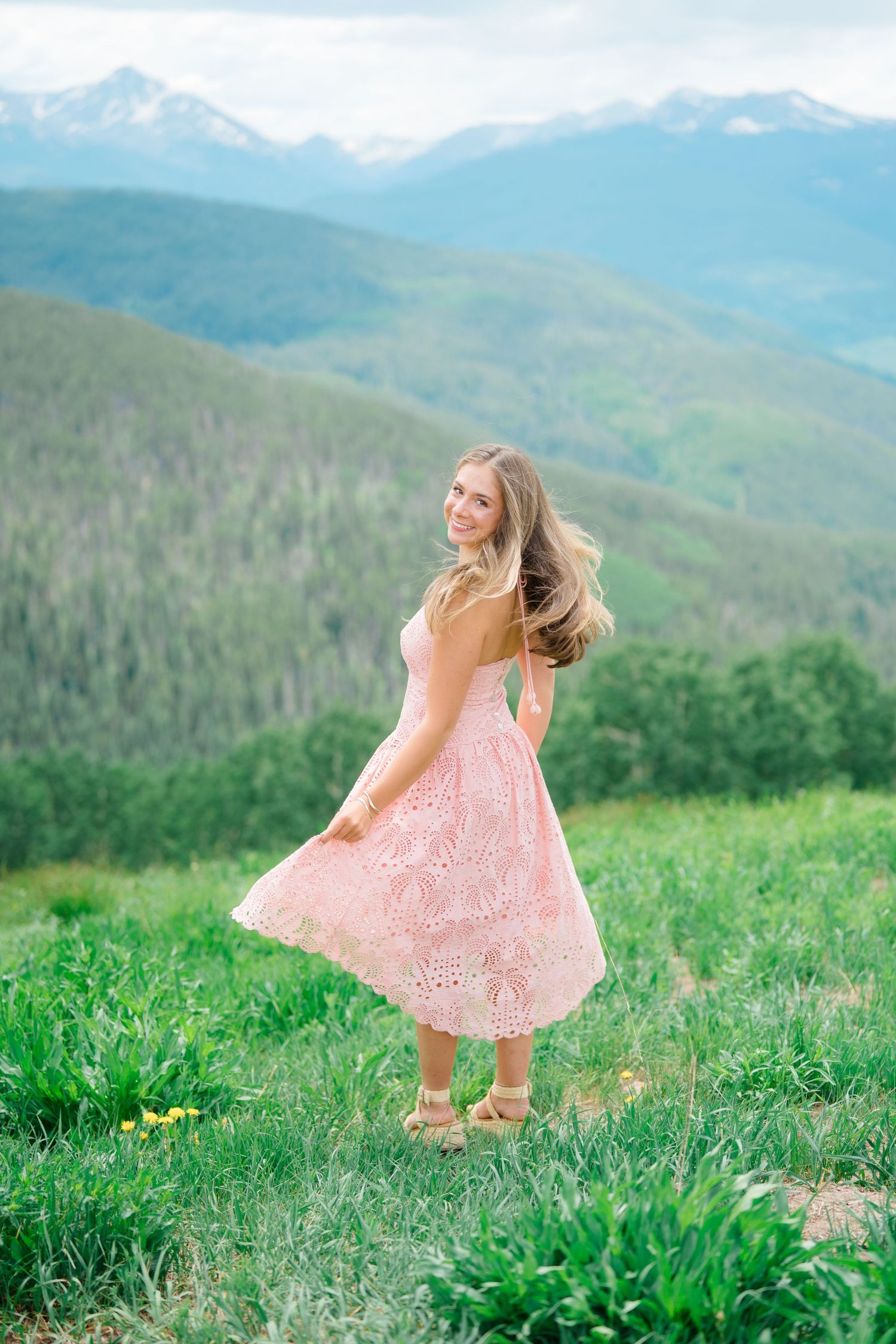 A high school senior woman in a pink lace dress twirls joyfully in a mountain meadow in vail with snow-capped peaks in the background.