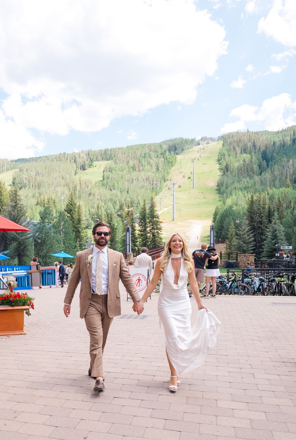 Bride and groom walking hand in hand at a mountain ski resort wedding venue on a sunny summer day.