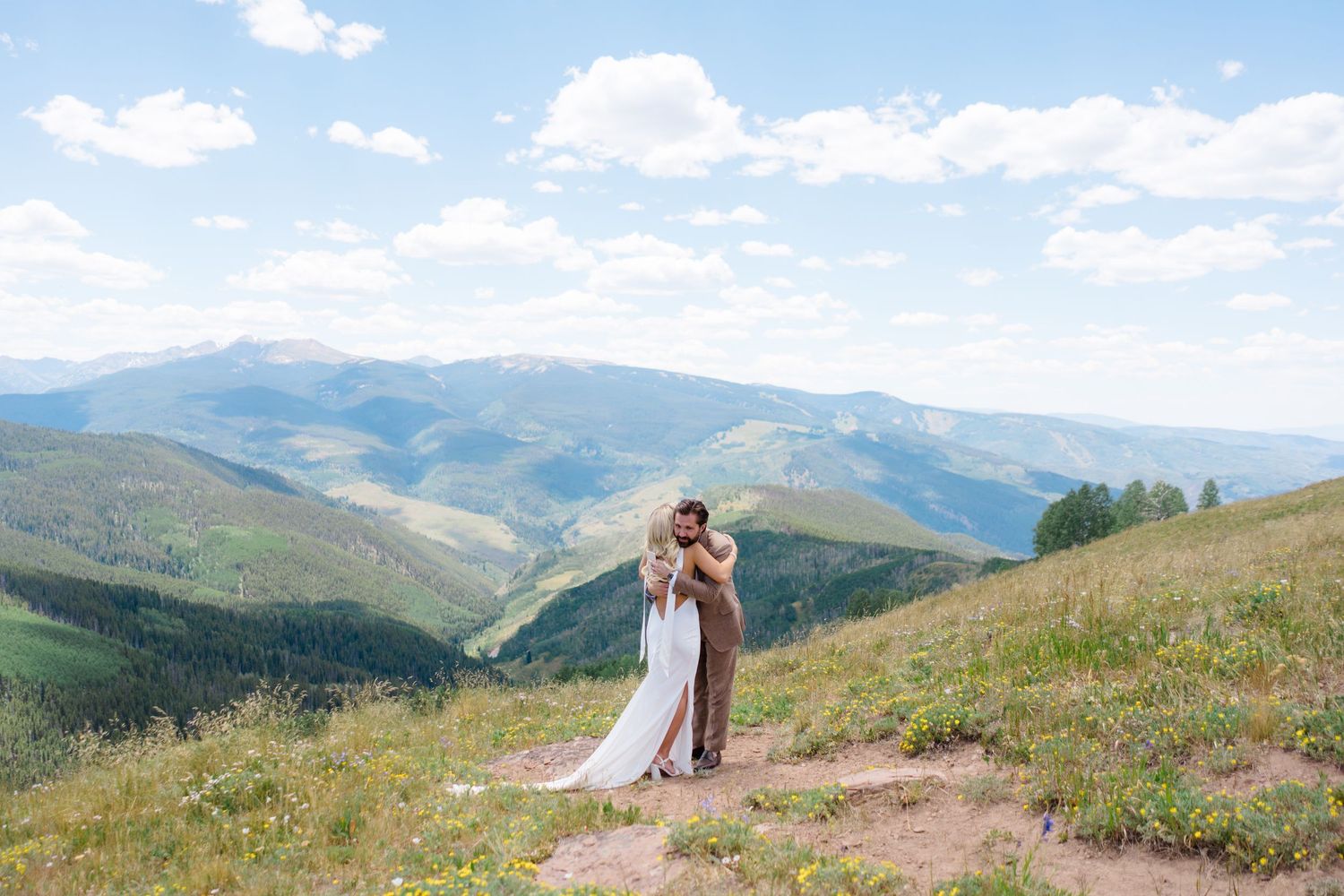 Bride and groom embracing on a mountain hillside with sweeping green valley views under a blue sky.