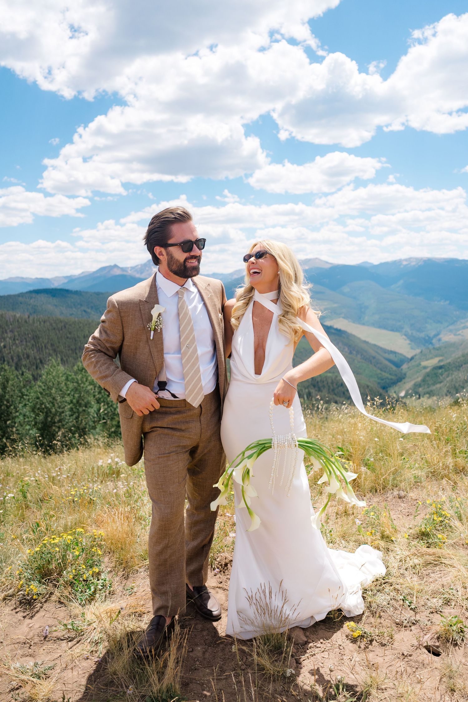 Bride and groom in sunglasses pose on a mountain hilltop, she in a white gown, he in a brown suit, with scenic peaks behind.