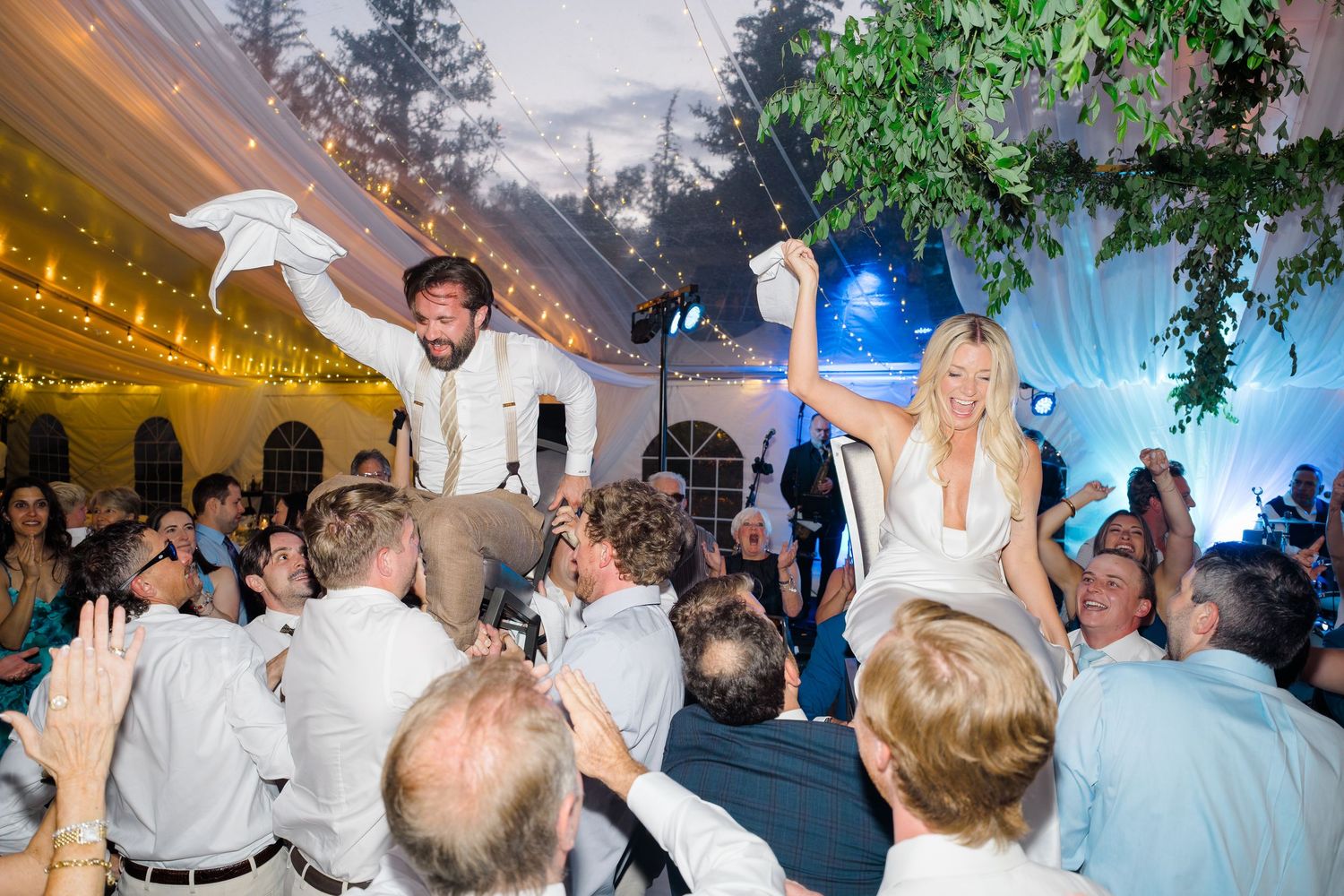 Bride and groom lifted on chairs during lively wedding reception dance celebration under festive tent lights.