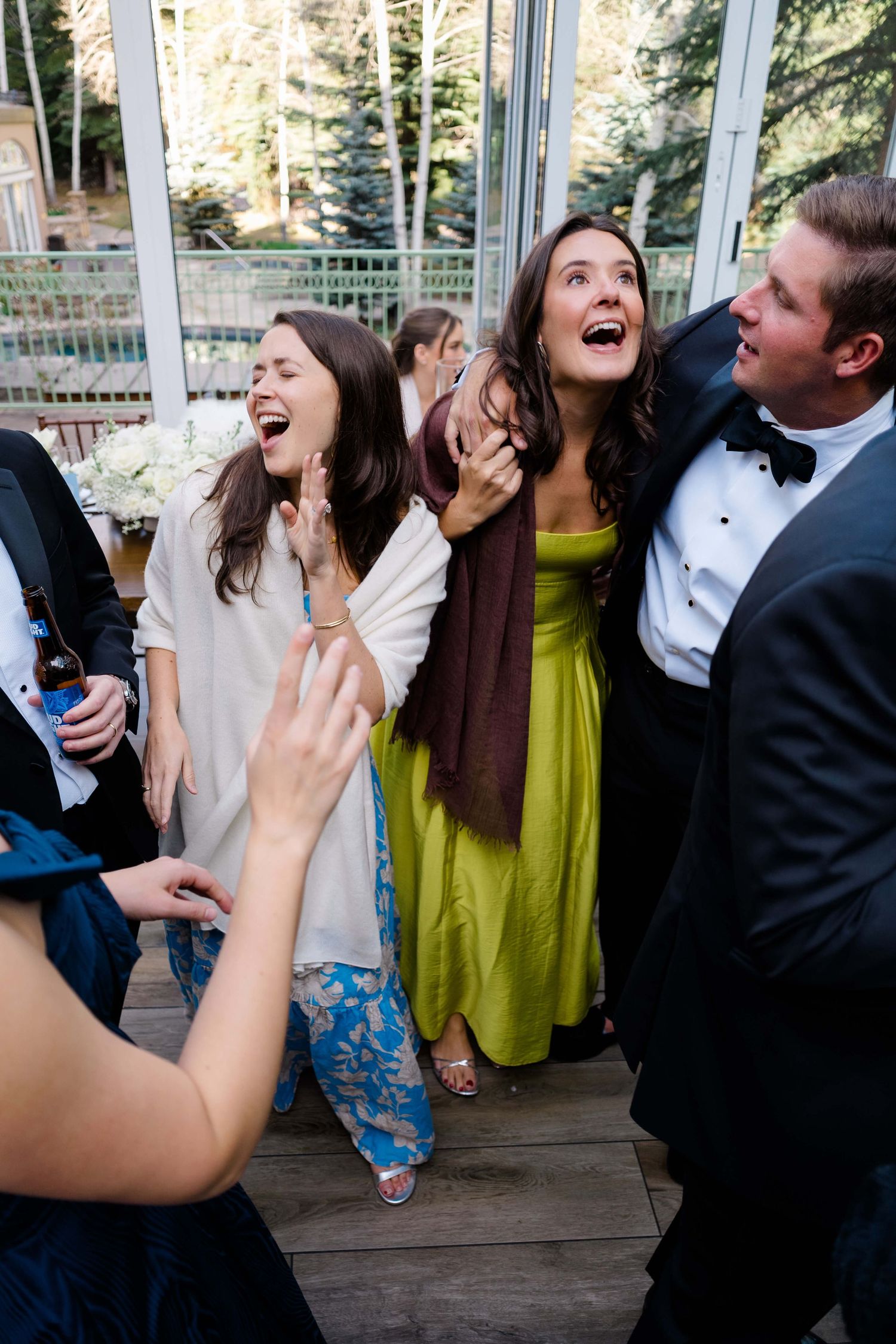 Wedding guests in formal attire laugh and celebrate joyfully on a wooden deck outside a glass venue.