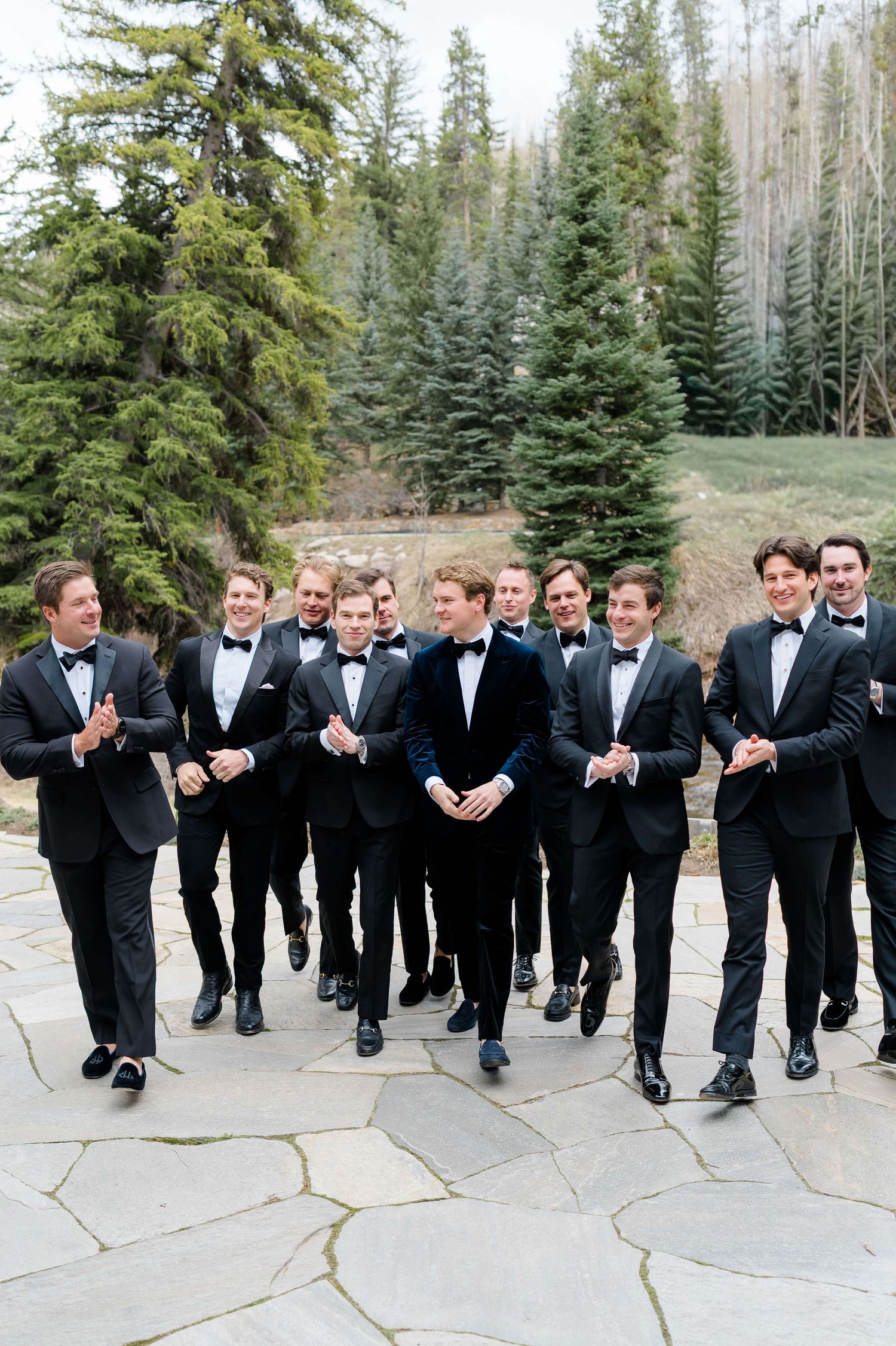 Groomsmen in black tuxedos and bow ties walk together outdoors on stone patio with evergreen trees in background.