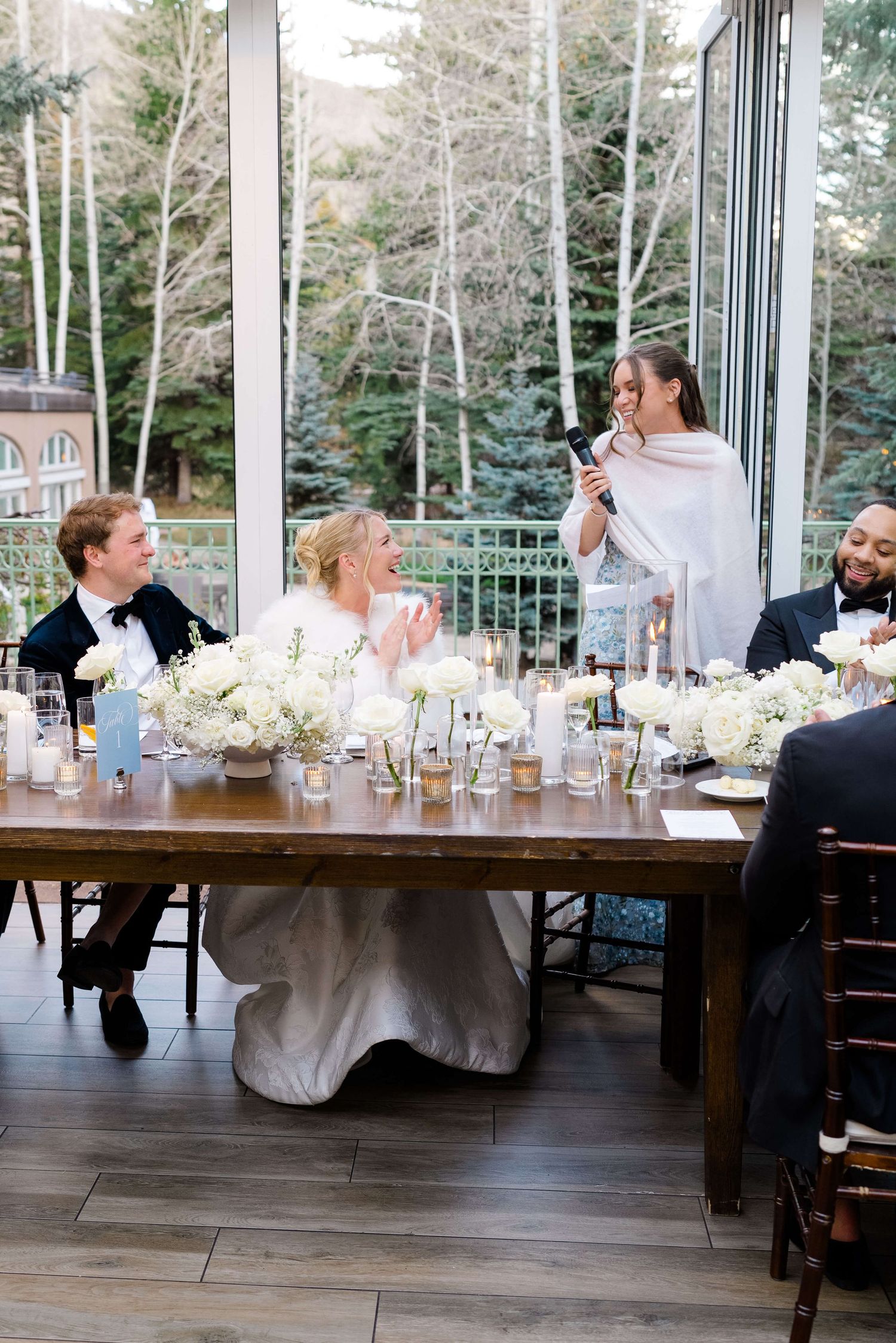 Bride and groom laugh during wedding reception toast at elegant wood table with white floral arrangements.