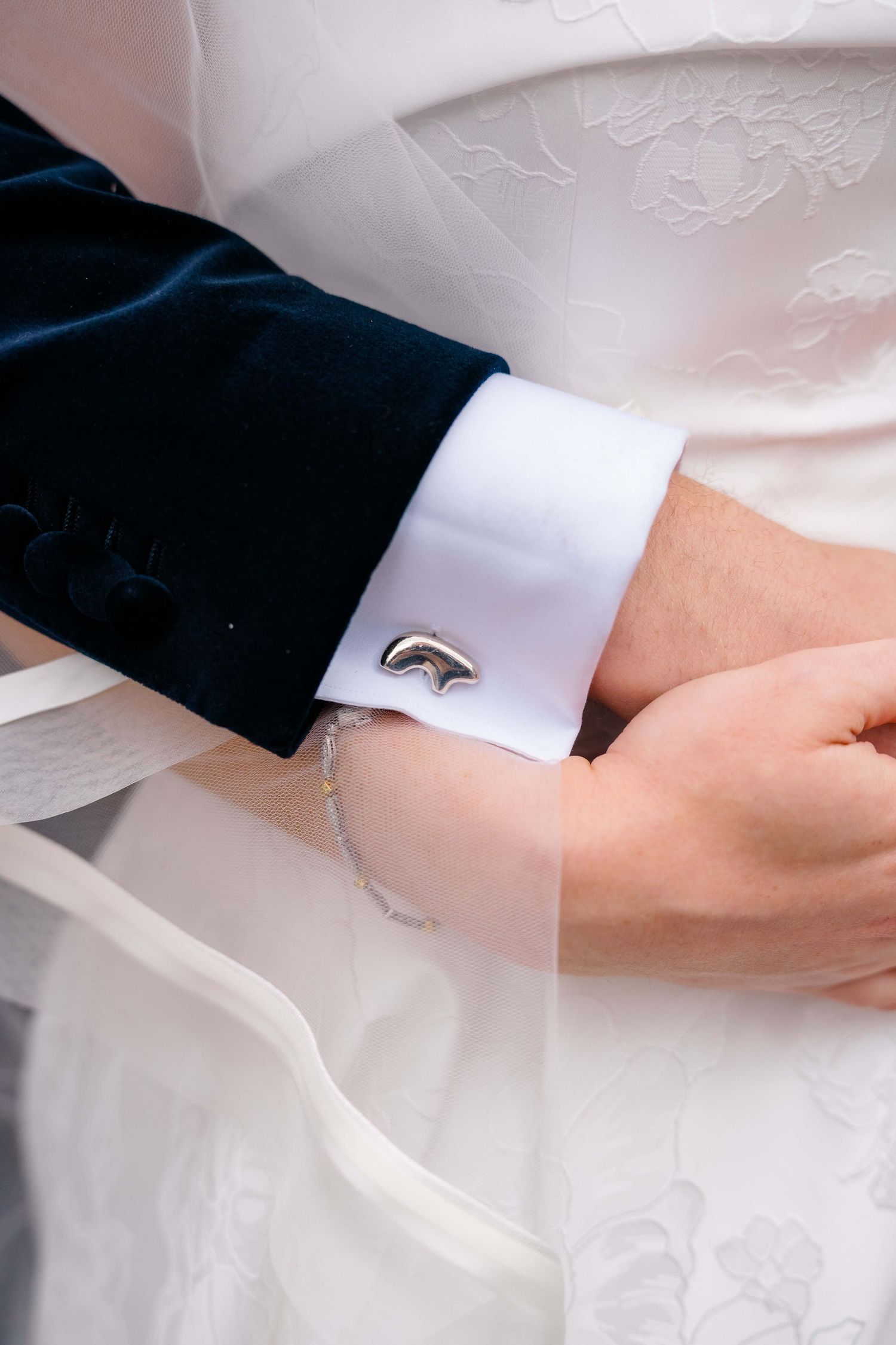 Close-up of groom's silver cufflink on white dress shirt sleeve beside bride's white wedding gown.