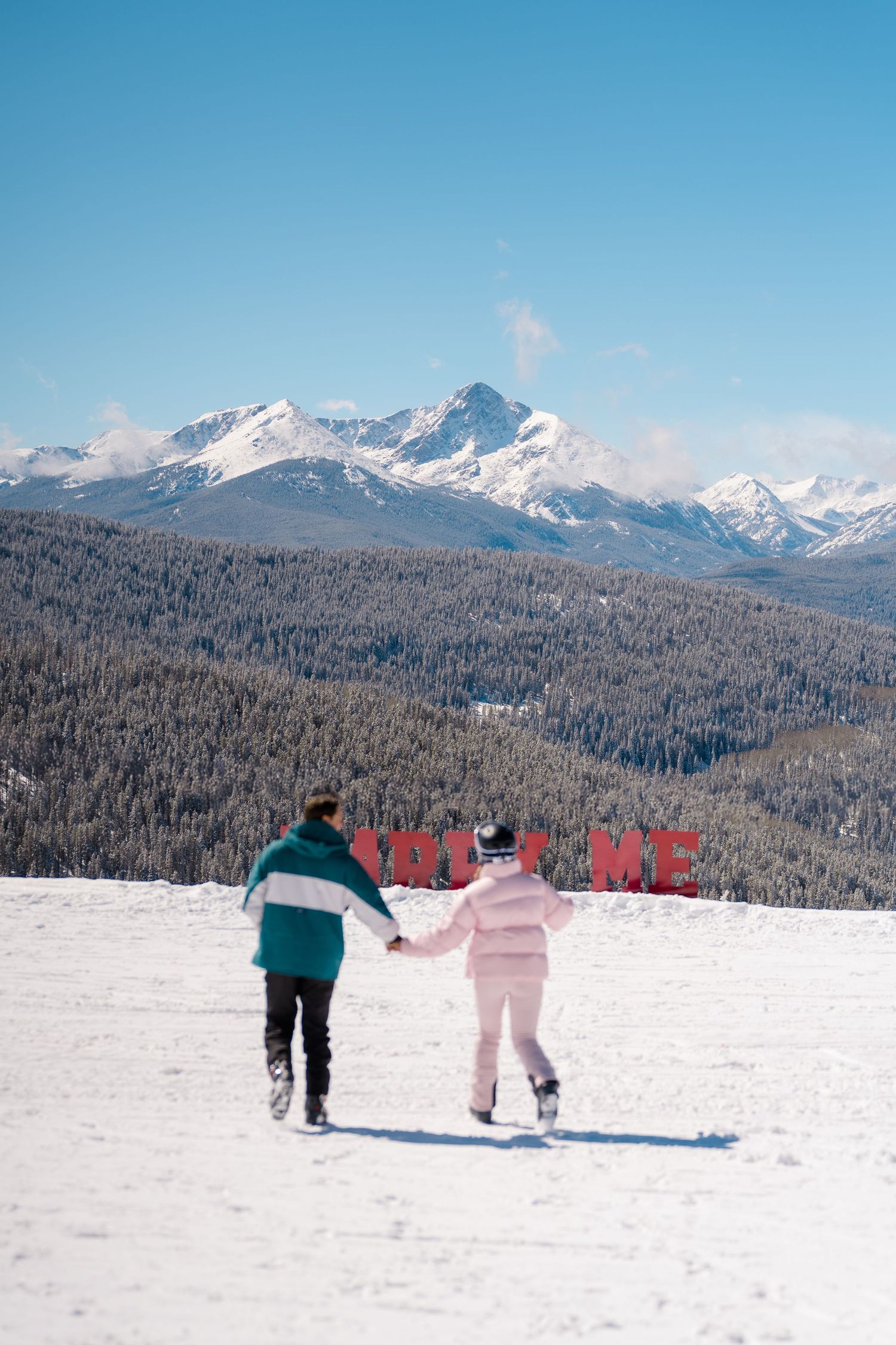 Skiers traverse a snowy mountain slope with majestic snow-capped peaks in the background under clear blue skies.