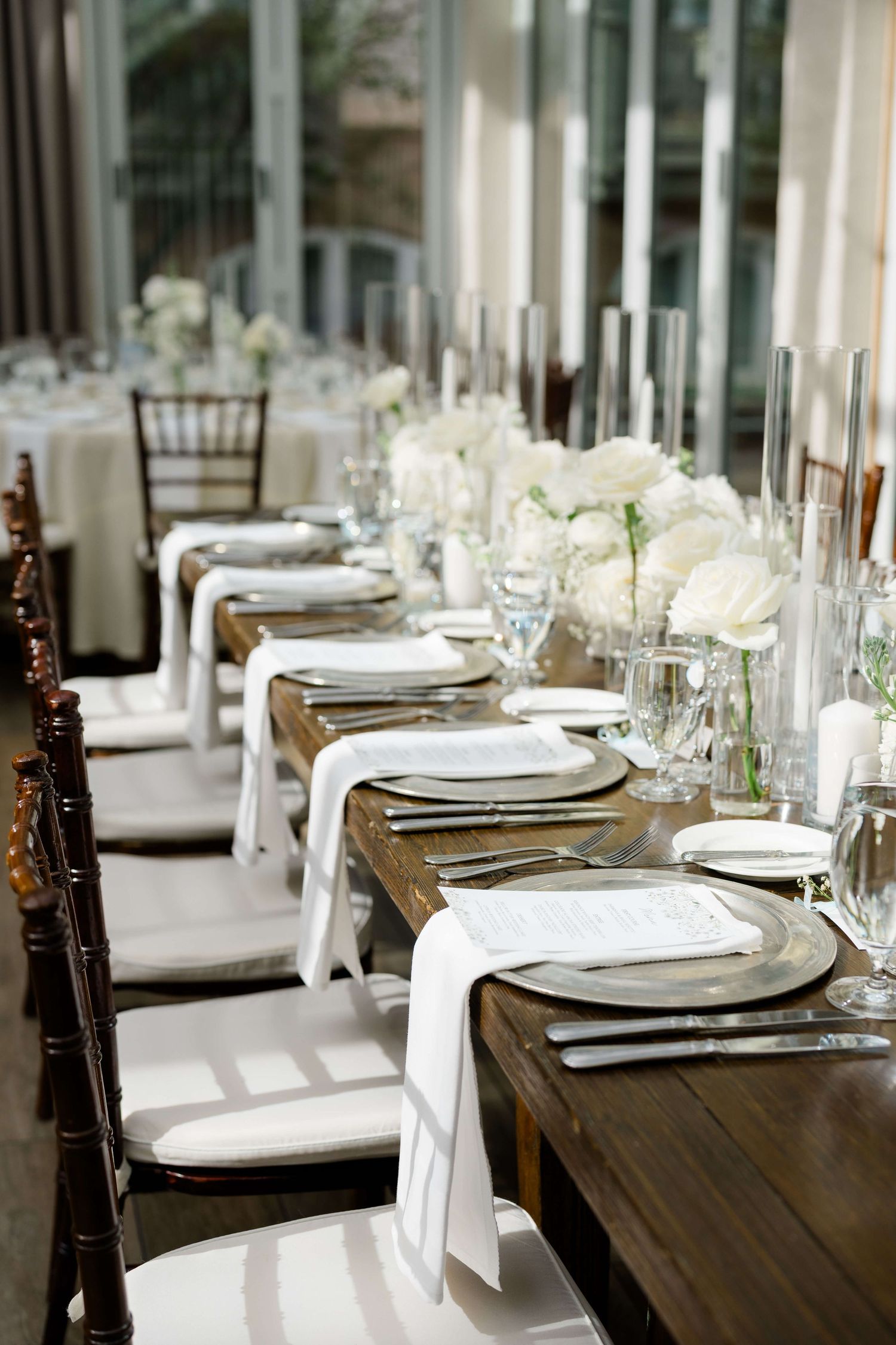 Elegant wedding reception table with white floral centerpieces, crystal glassware, and white napkins on a rustic wooden table.