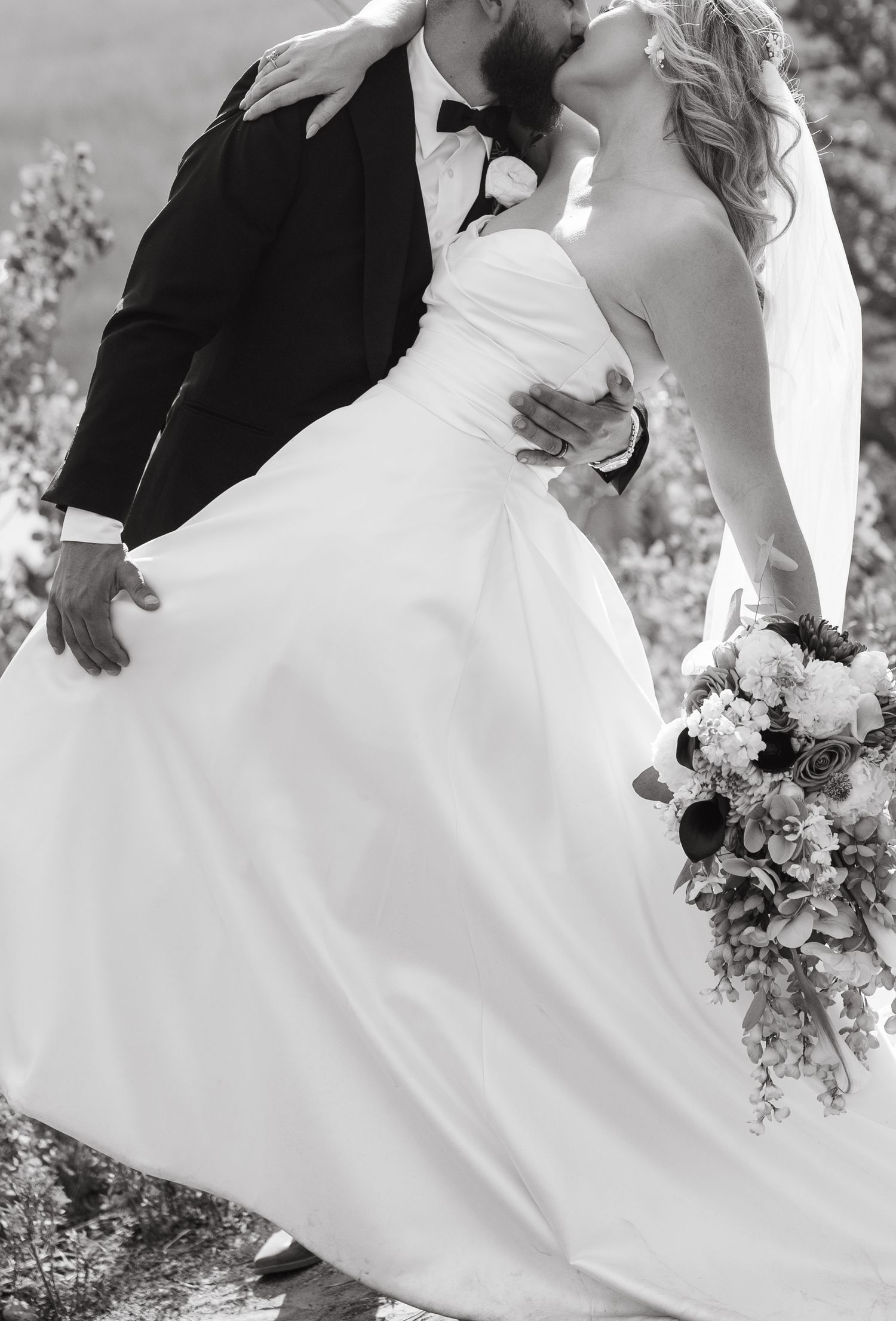 A romantic black and white wedding photo showing a couple kissing while holding a floral bouquet in an outdoor setting.