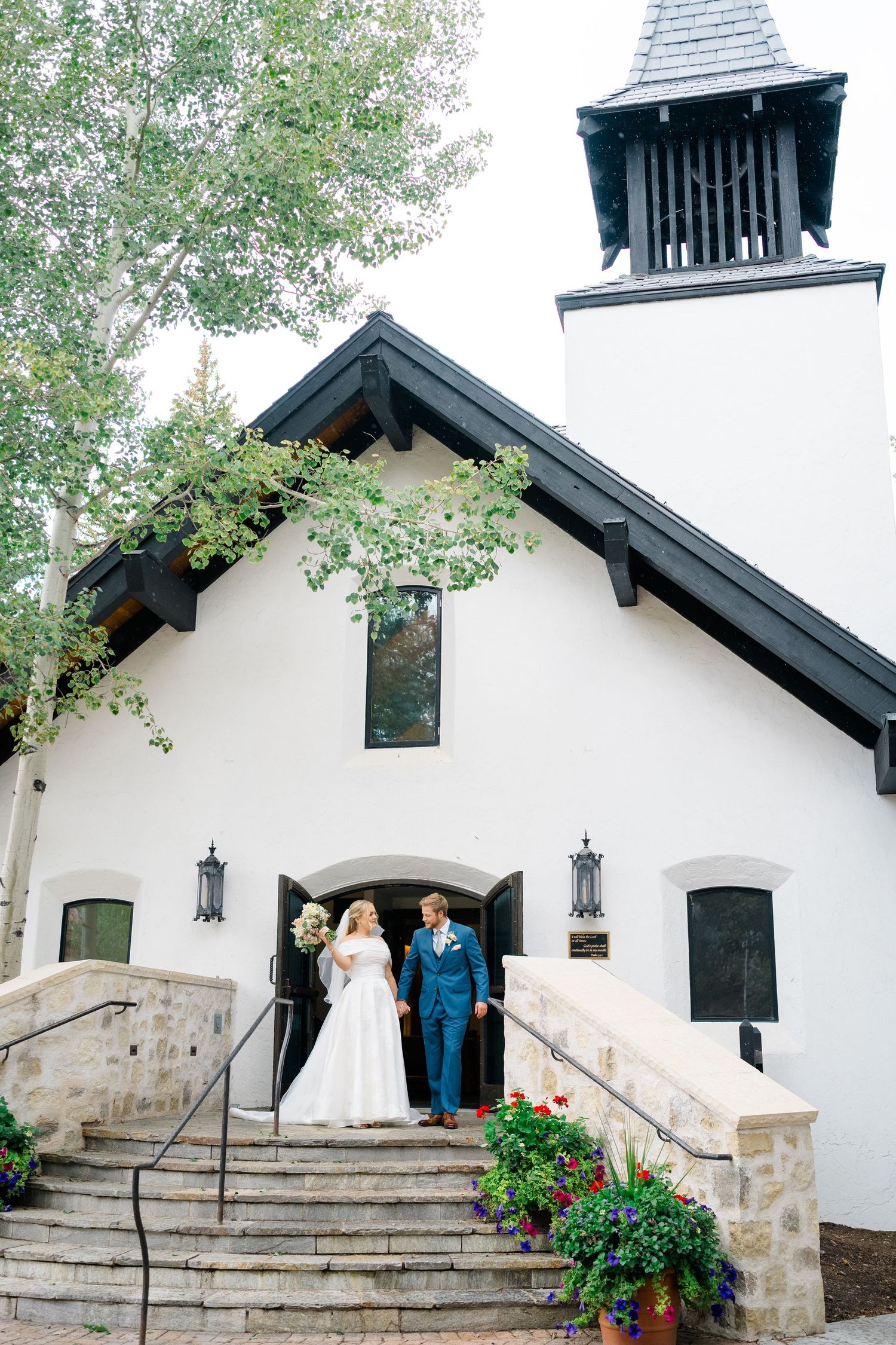A white chapel with stone steps and black trim features vibrant red flowers in planters along the entrance during vail wedding.