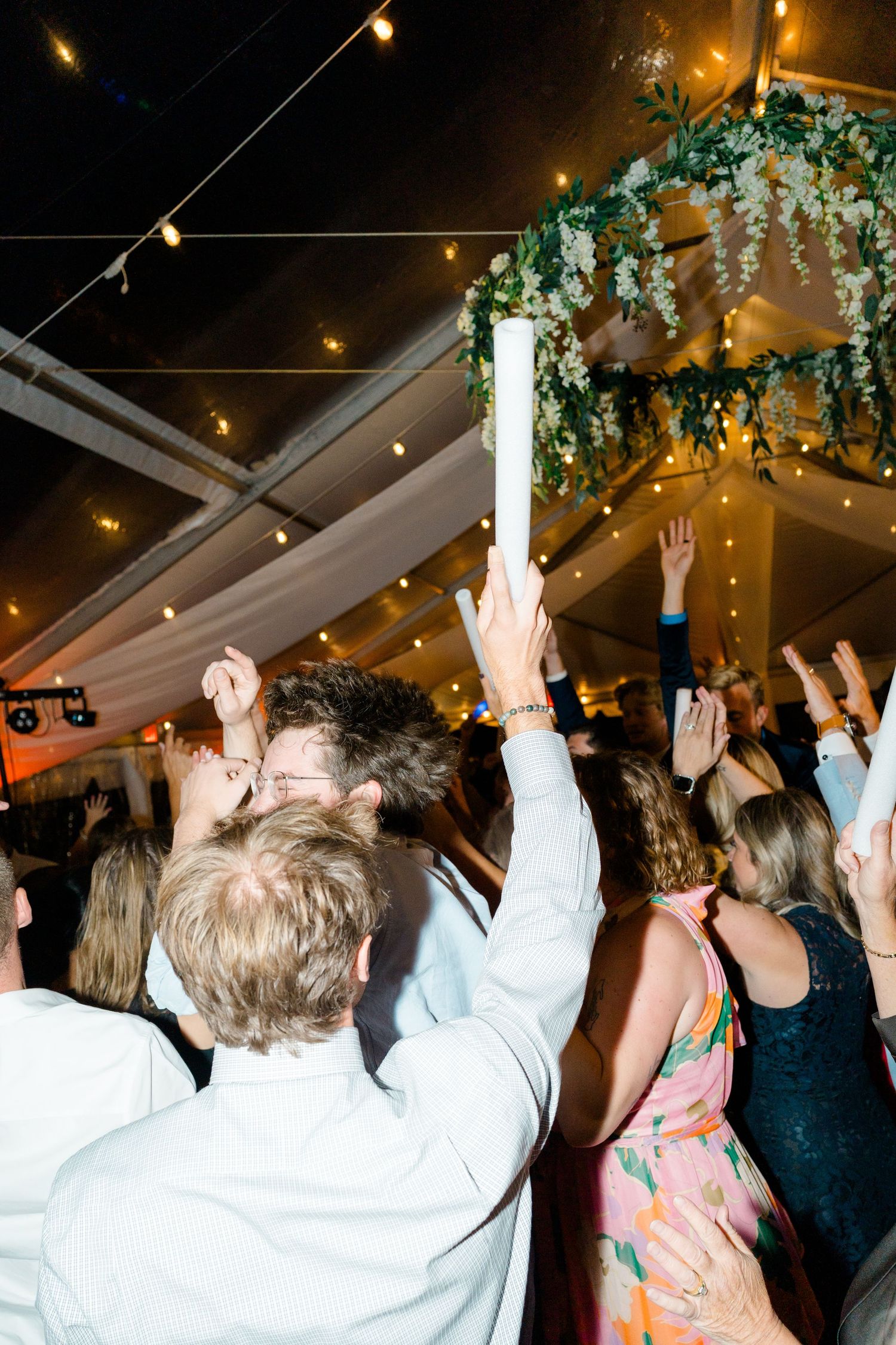 Party guests raise glowing foam sticks in celebration under string lights and hanging greenery at an indoor event.