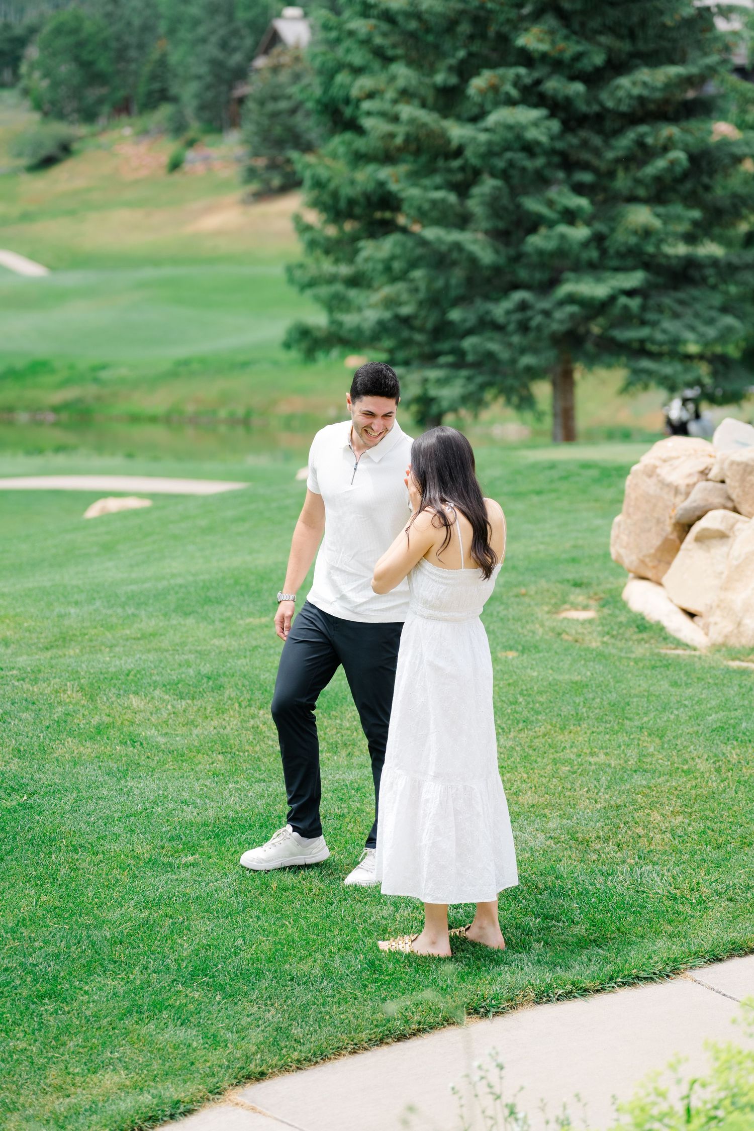 A couple in white outfits walks together on a grassy hillside with evergreen trees in the background.