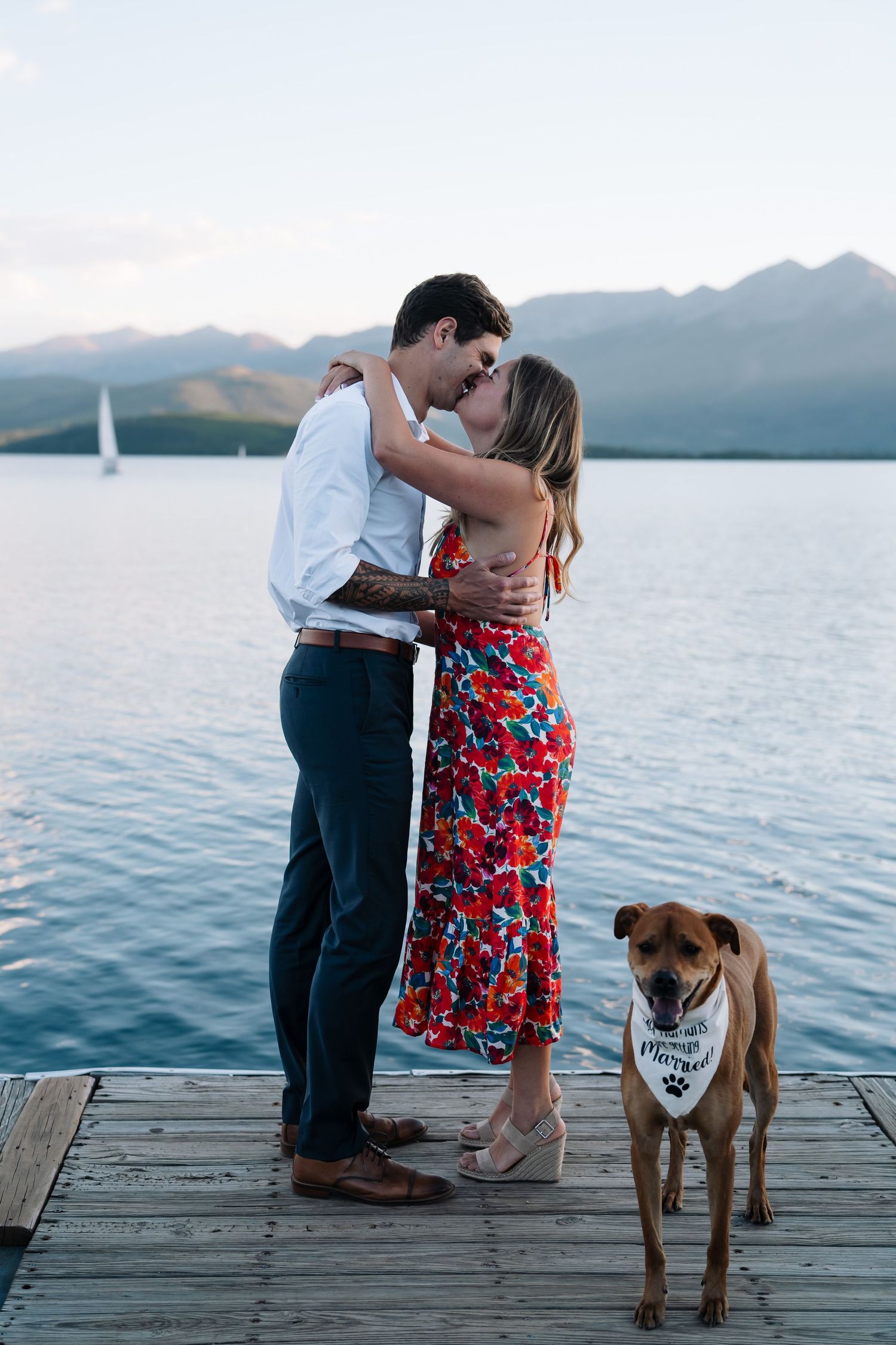 A couple shares a romantic moment on a lakeside dock with their brown dog wearing a bandana against a mountain backdrop.