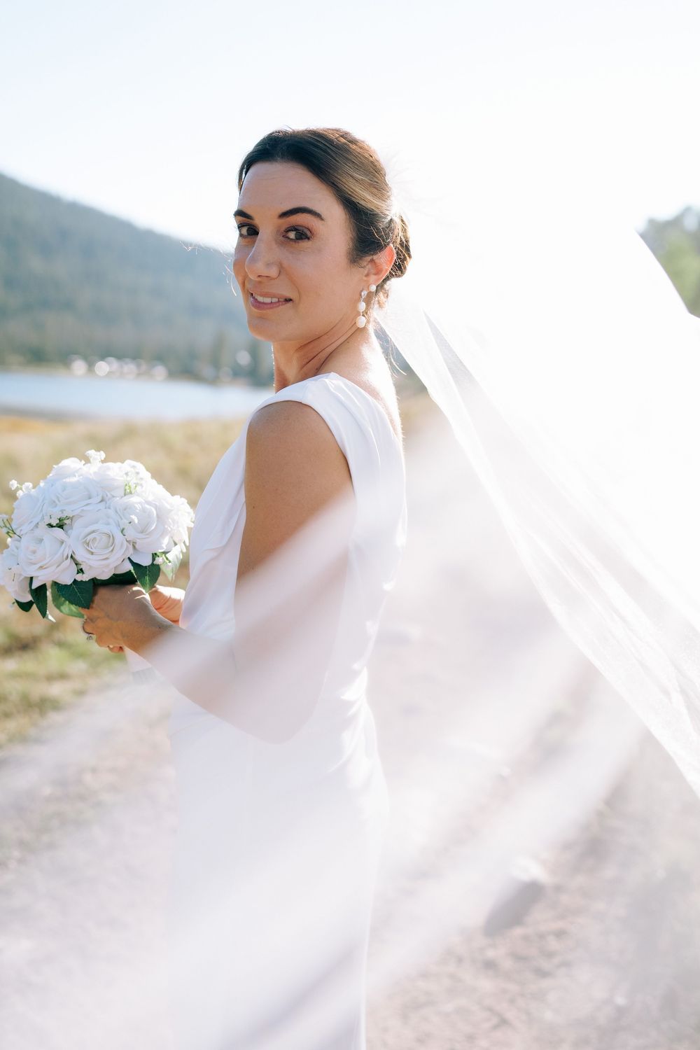 Bride in white gown and veil holds rose bouquet outdoors, smiling over shoulder with sunlit lake and mountains behind her.