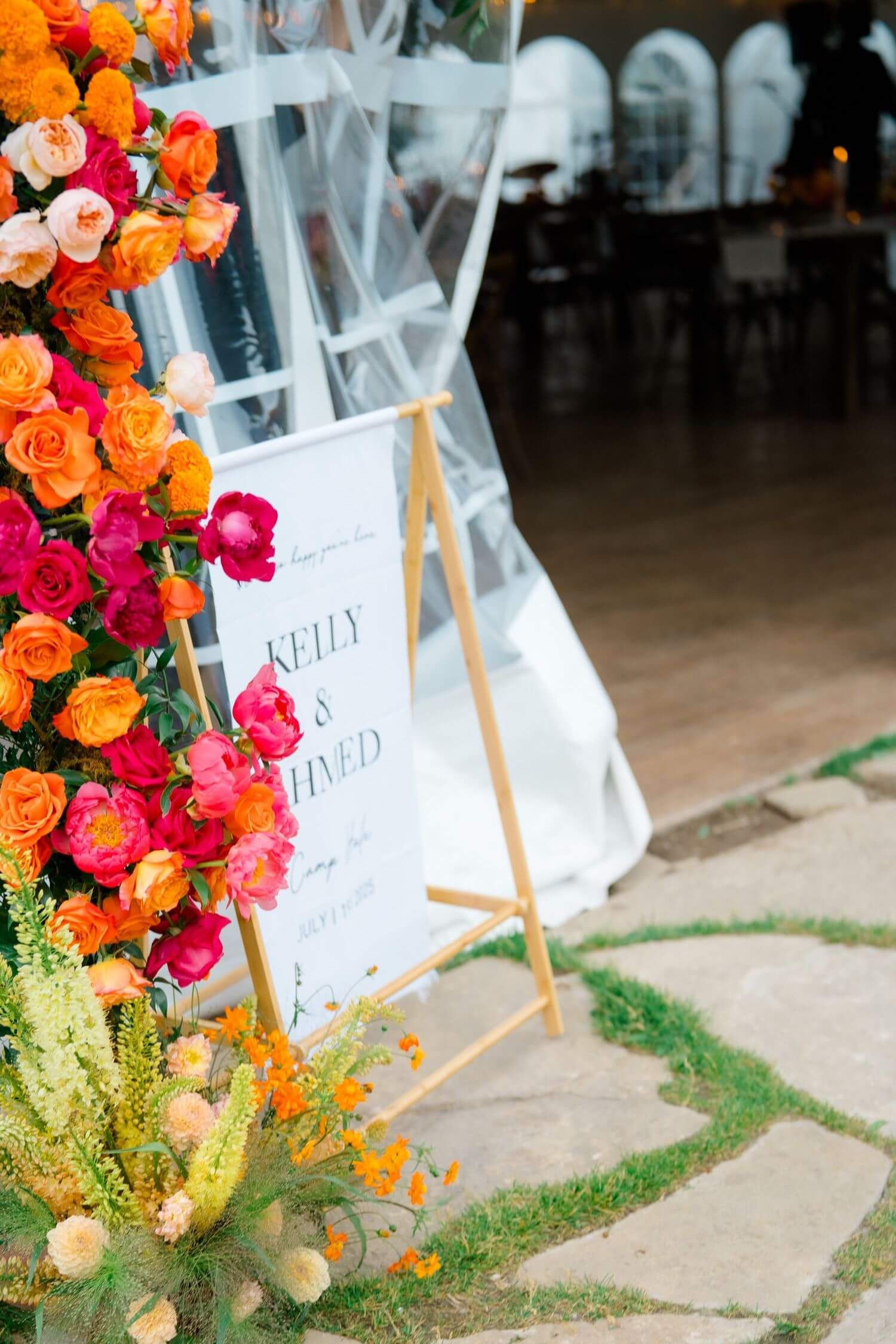 Vibrant orange and pink floral arrangements with greenery decorate white seating signs at an outdoor wedding venue at camp hale wedding