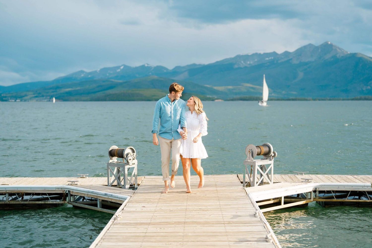 A couple walks along a wooden dock extending into a scenic mountain lake with sailboats visible in the distance at lake dillon.