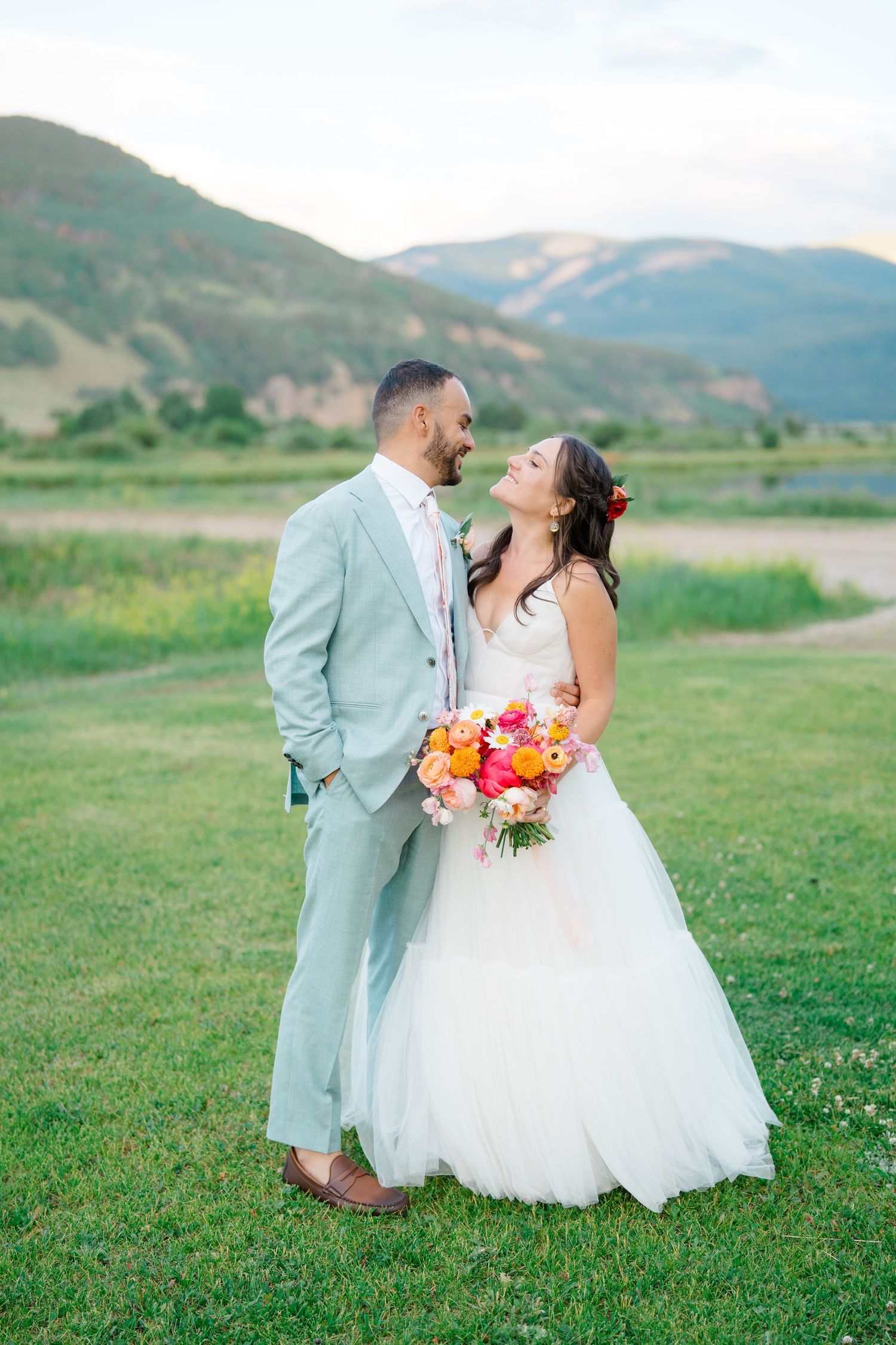 A couple shares a romantic moment in a grassy meadow with mountain views and colorful wedding bouquet.