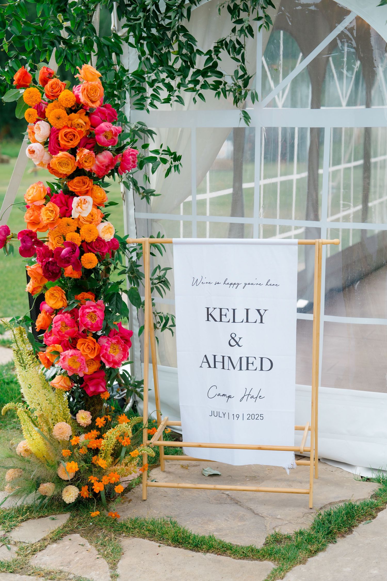 Vibrant orange and pink floral wedding decorations with signage displayed along a white greenhouse wall.