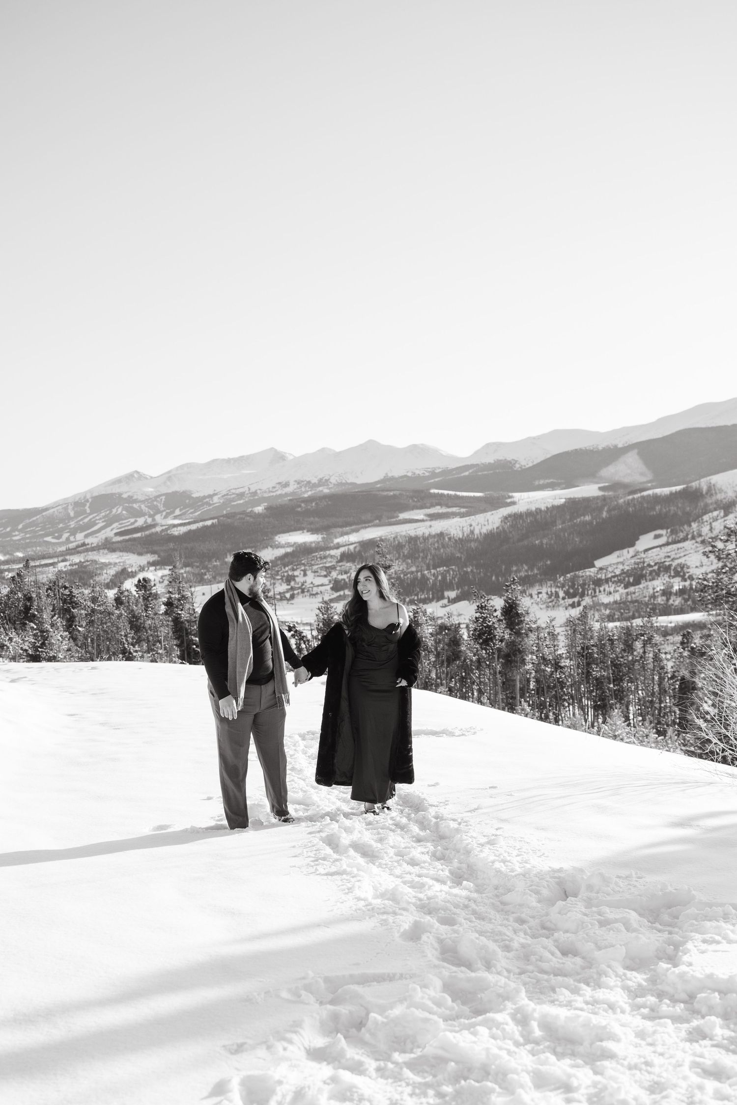 A couple stands together in a snowy mountain landscape with snow-capped peaks and evergreen forests in the distance.