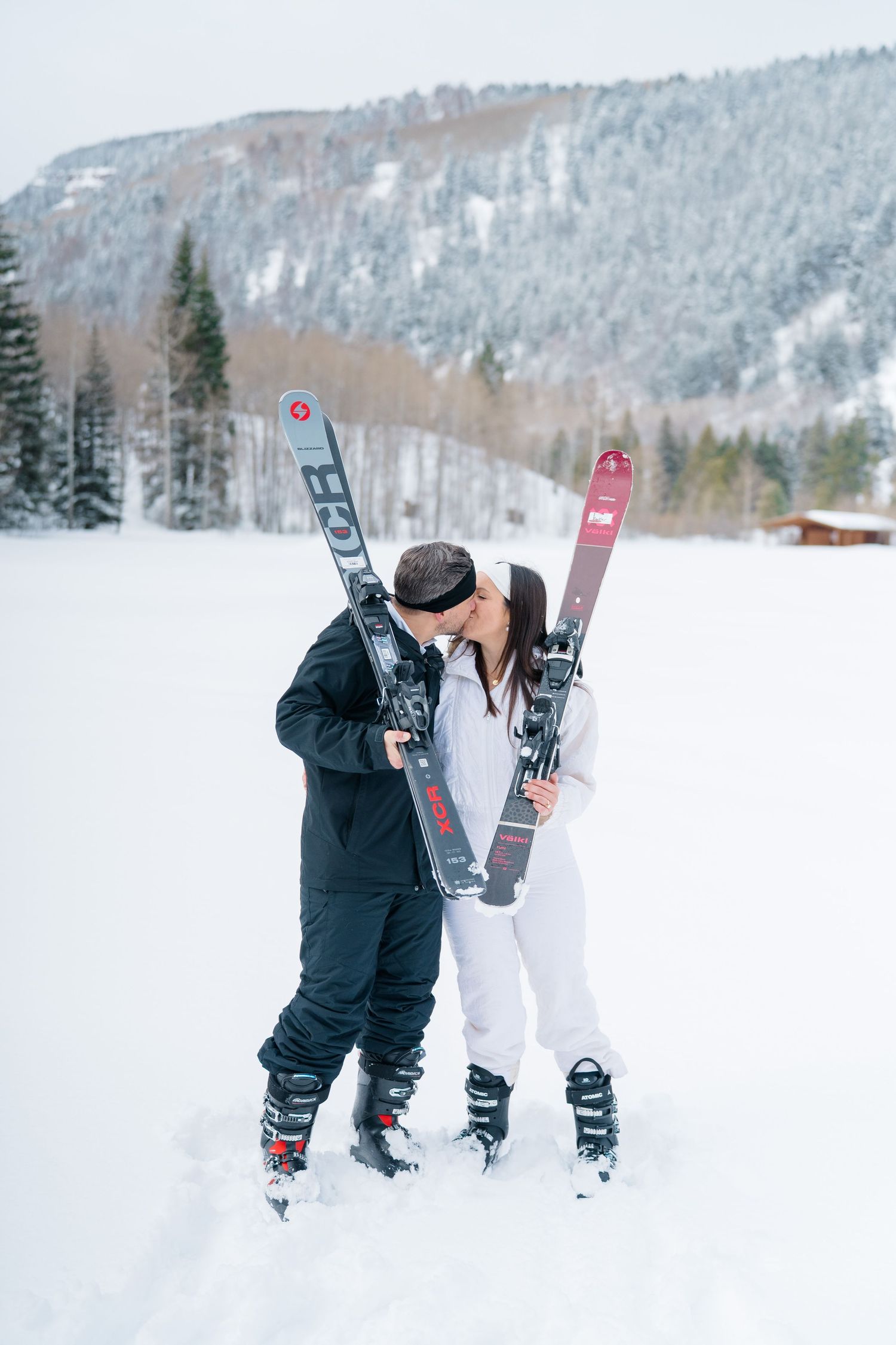 Skiers in black winter gear prepare their equipment on a snowy mountain slope with snow-covered trees in the background.