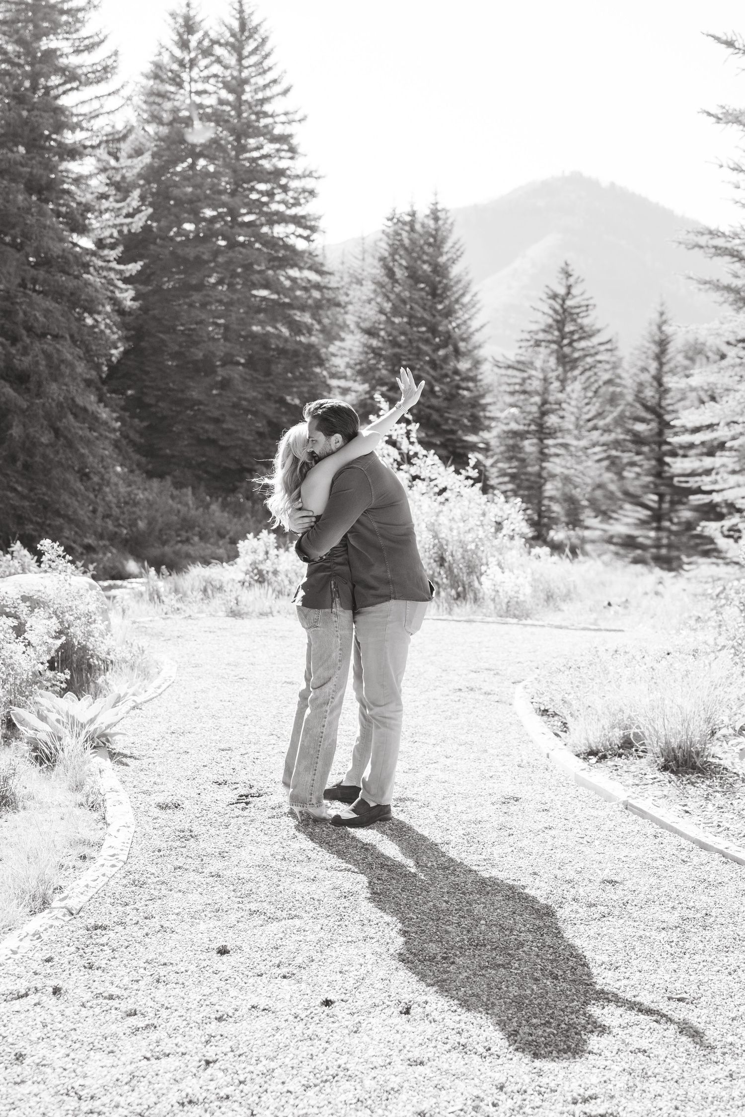A couple shares a romantic embrace on a snowy mountain trail surrounded by evergreen trees and winter scenery.