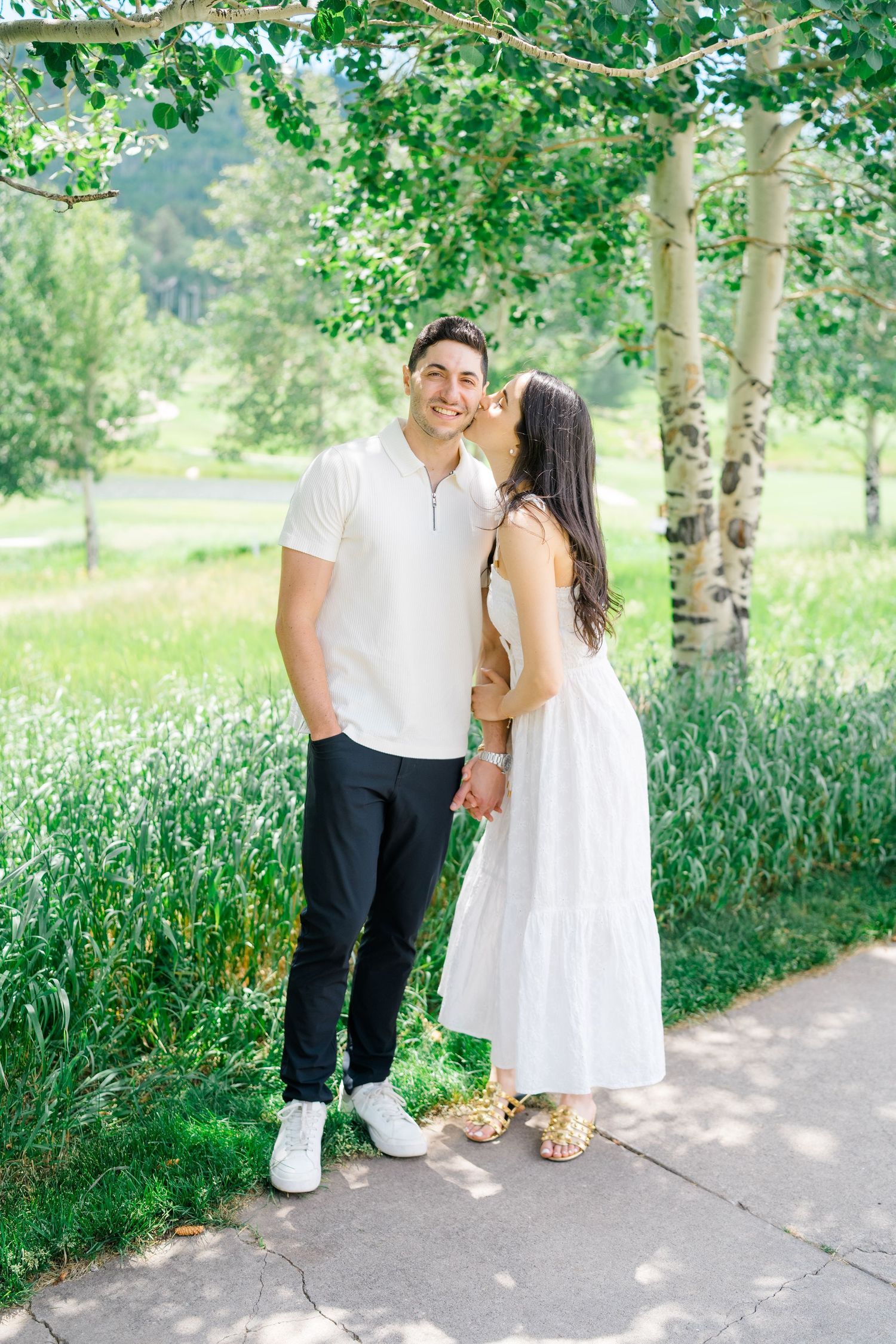 A couple walks hand in hand along a tree-lined path in coordinating white outfits on a sunny day.
