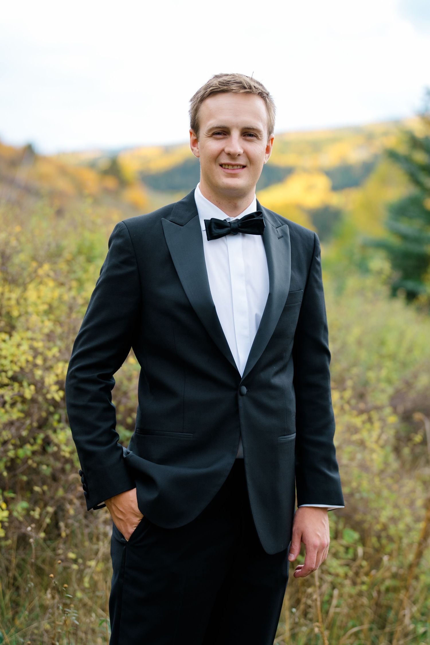 Groom in classic black tuxedo and white bow tie poses in outdoor autumn setting with golden foliage.