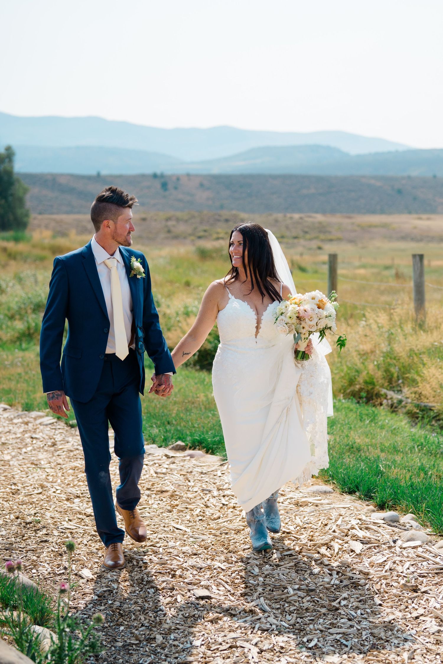 A couple in wedding attire walks along a scenic mountain path with blue skies and rolling hills in the background.